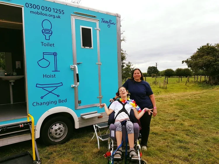 A young girl named Tamsin is in a wheelchair and a woman with her are smiling in front of a blue mobile bathroom truck labeled "Tamsin" with signs indicating it has a toilet, hoist, and changing bed. The truck is parked on a grassy field.