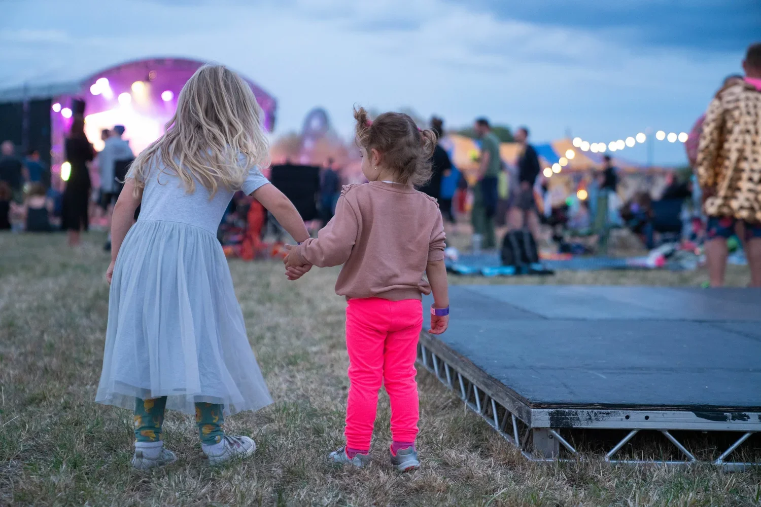 Two young girls holding hands at an outdoor concert during dusk, with a stage lit with pink and purple lights in the background.