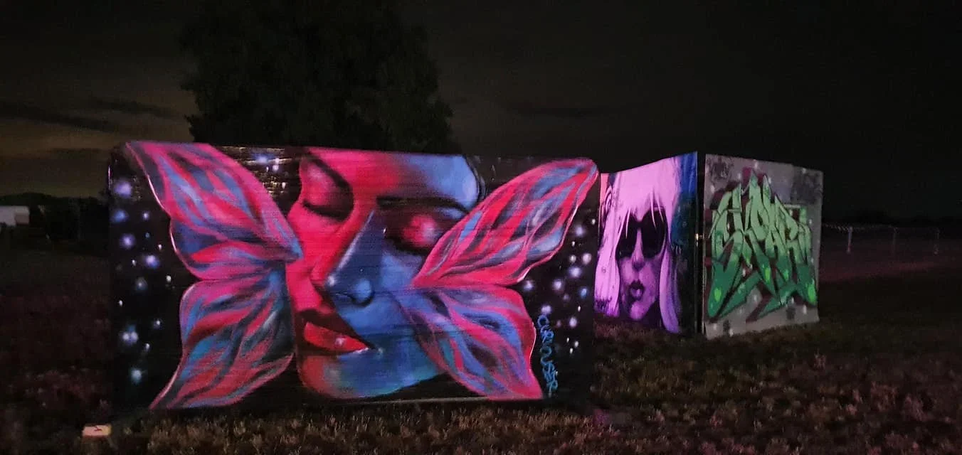 Nighttime outdoor view of colorful graffiti art on three large panels, including a butterfly with a human face, a woman's face with sunglasses and purple hair, and green abstract graffiti.