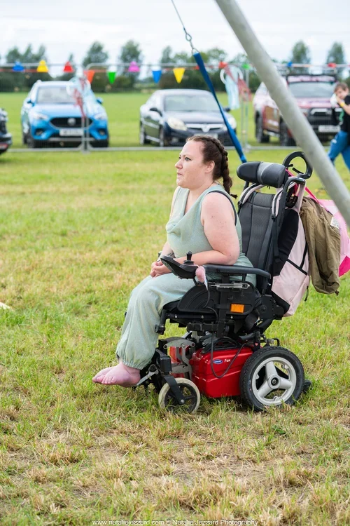 A woman in a motorized wheelchair sitting on grass near parked cars at an outdoor event about to partake in arial acrobatics.