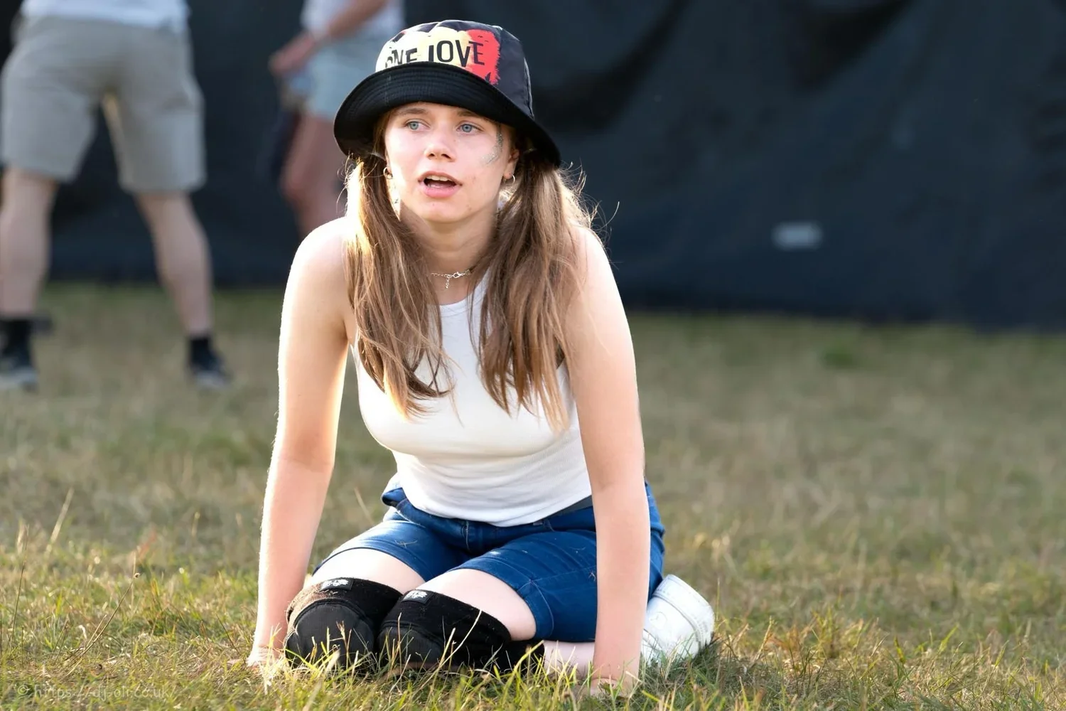 Young woman with long hair wearing a white tank top, denim shorts, kneeling on grass with hands on the ground, looking happy and free, wearing a black bucket hat with red and white logo, black knee pads, and white sneakers.