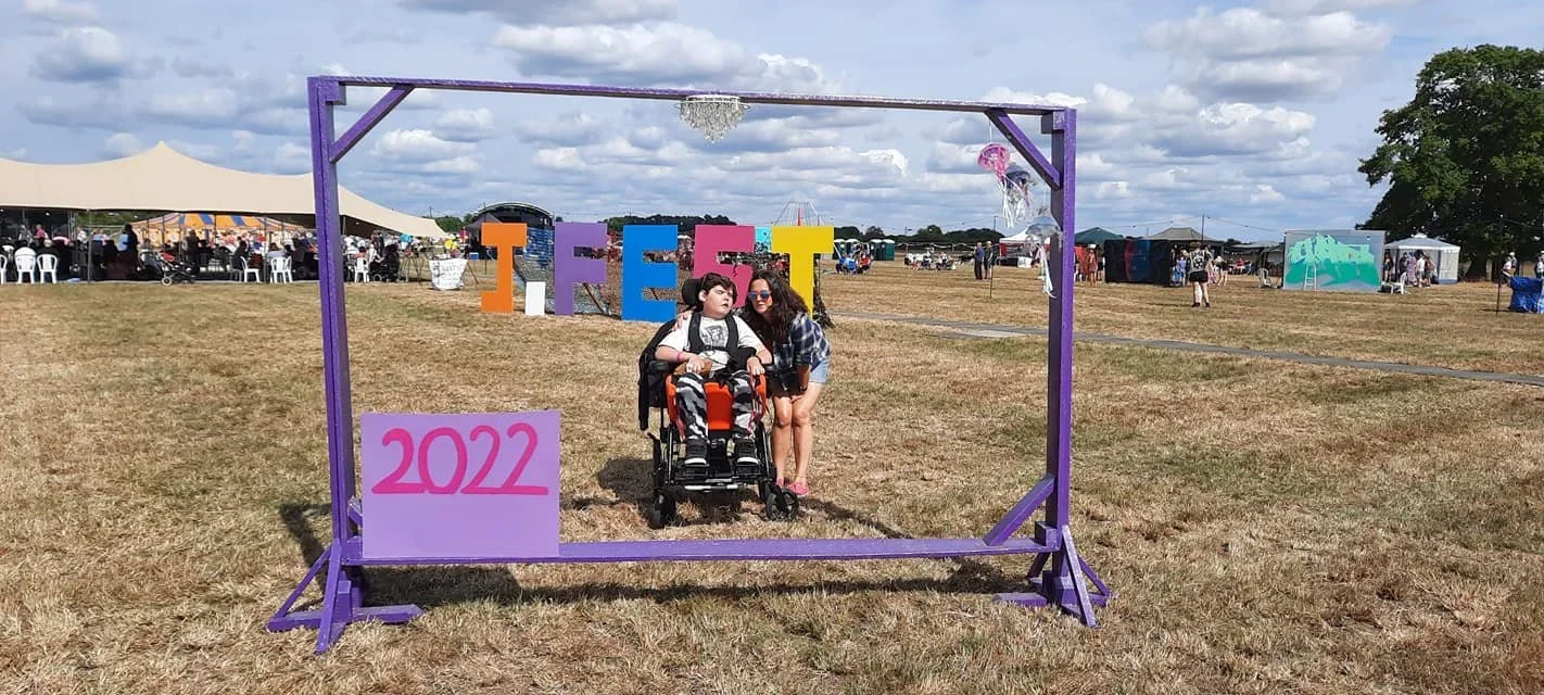 Two young people, one in a wheelchair, posing for a photo under a purple frame with the word 'J-FEST' on it, at an outdoor festival.