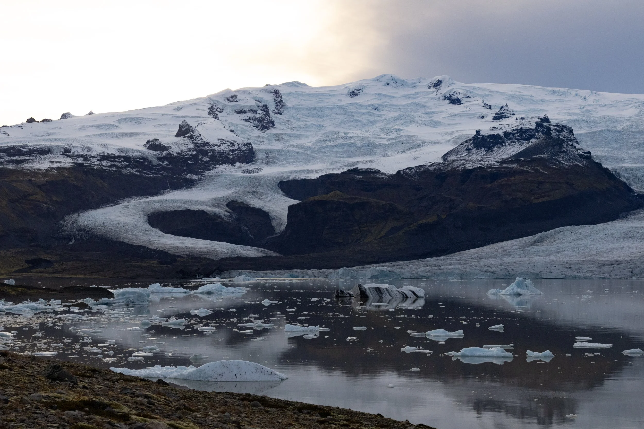 A glacier melting into a calm body of water with icebergs, with snow-capped mountains in the background on a cloudy day.