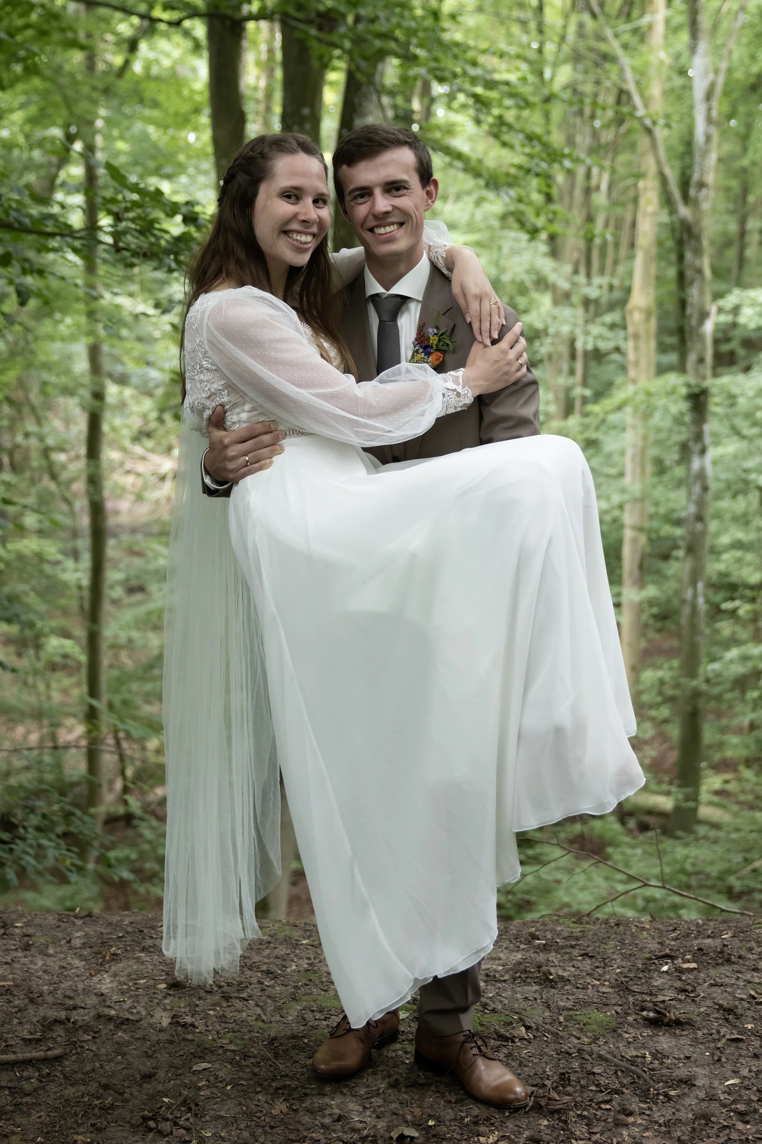 A couple dressed in wedding attire, with the bride in a white gown and the groom in a brown suit, standing in a forested area, with the groom carrying the bride in his arms, both smiling.