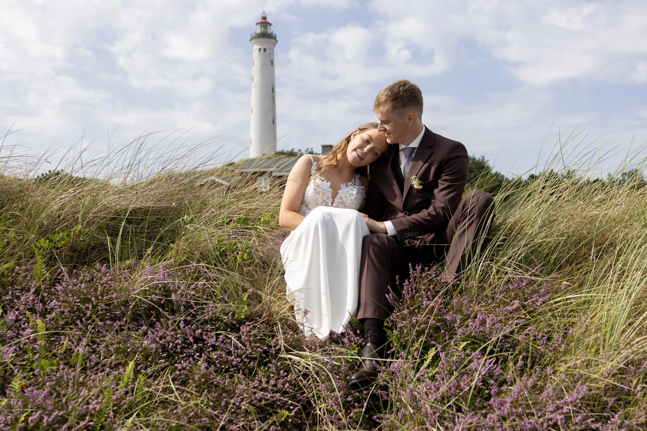 A newlywed couple in wedding attire sitting in a field of purple wildflowers with a lighthouse in the background under a partly cloudy sky.