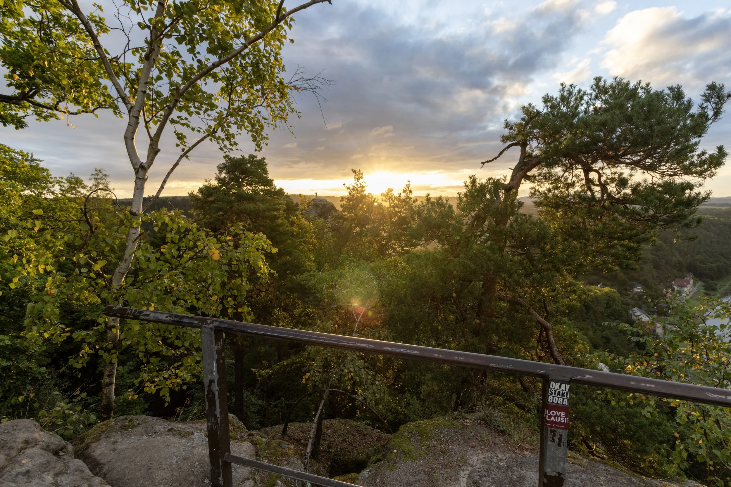 Sun setting over a lush green landscape with trees and houses visible in the distance. A metal railing with stickers is in the foreground.