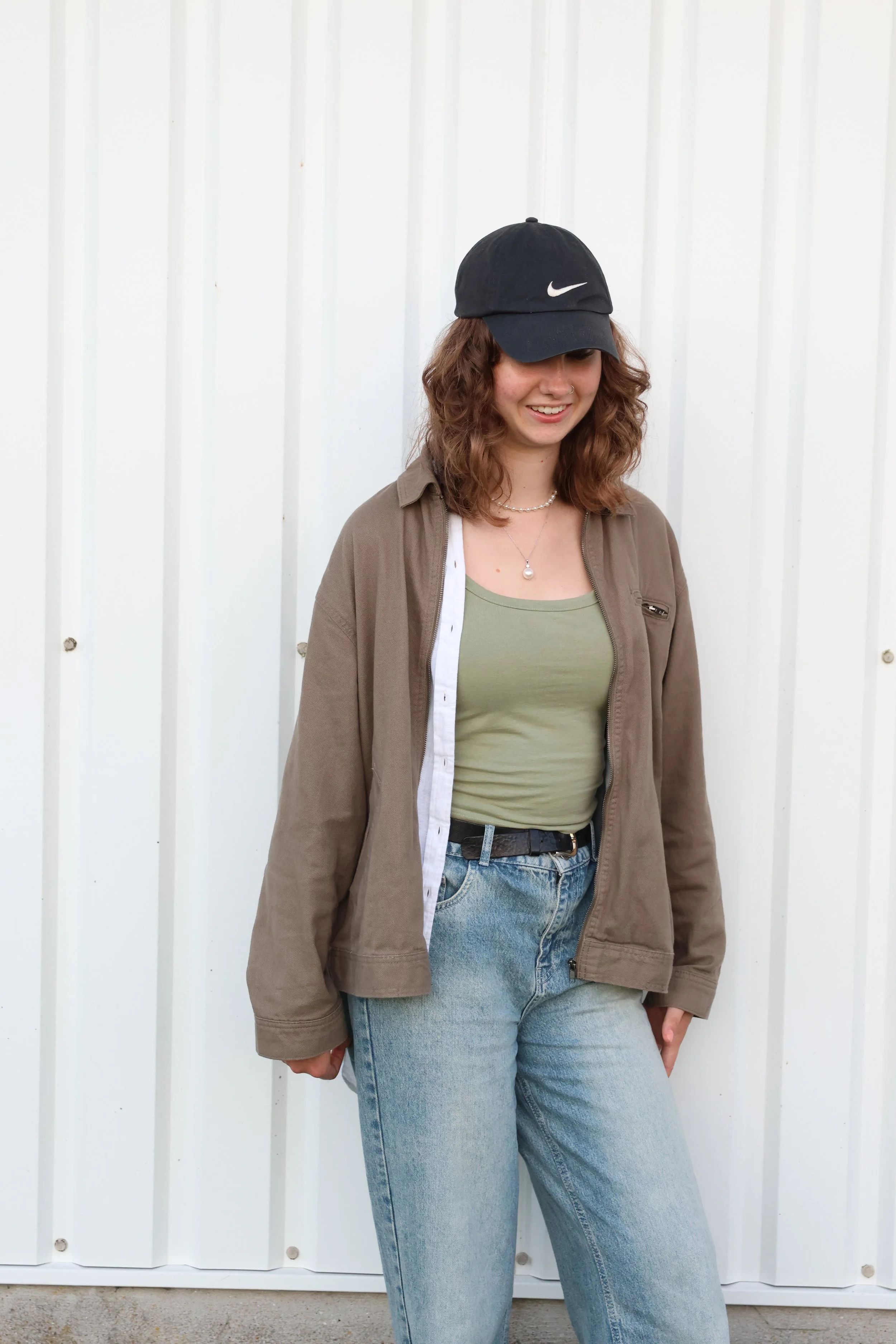 A young woman standing against a white metal wall, wearing a black Nike baseball cap, brown jacket over a green tank top, light blue jeans, and jewelry including a necklace and nose ring.