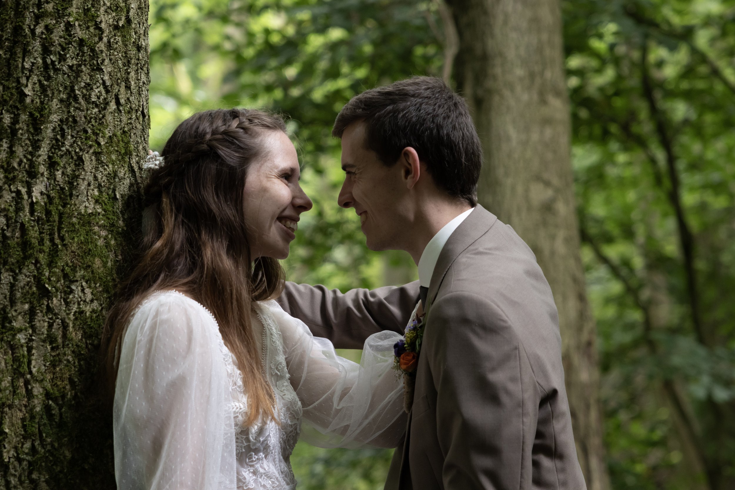 A couple in wedding attire exchange smiles in a forest, standing close with their foreheads nearly touching, surrounded by trees and green leaves.