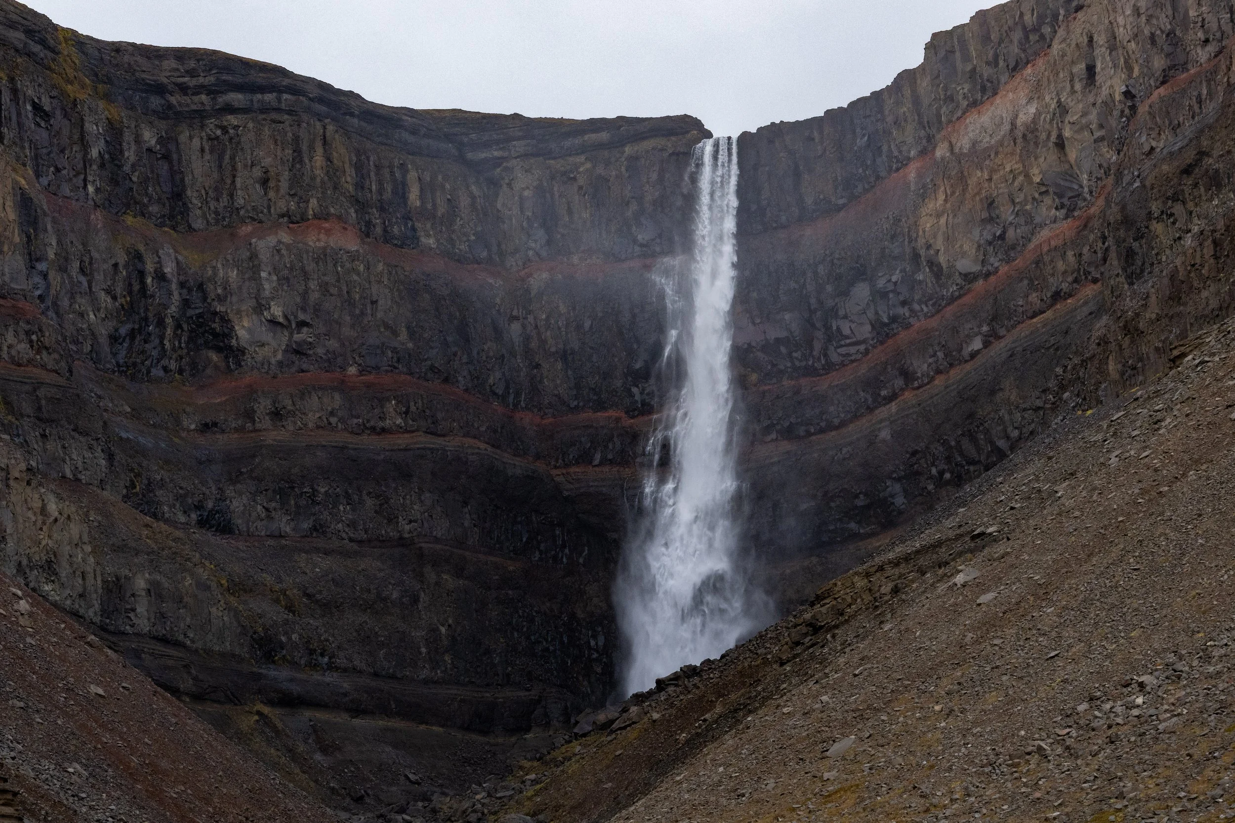 A tall waterfall cascading down a rugged, dark mountain cliff with a wide, rocky foreground.