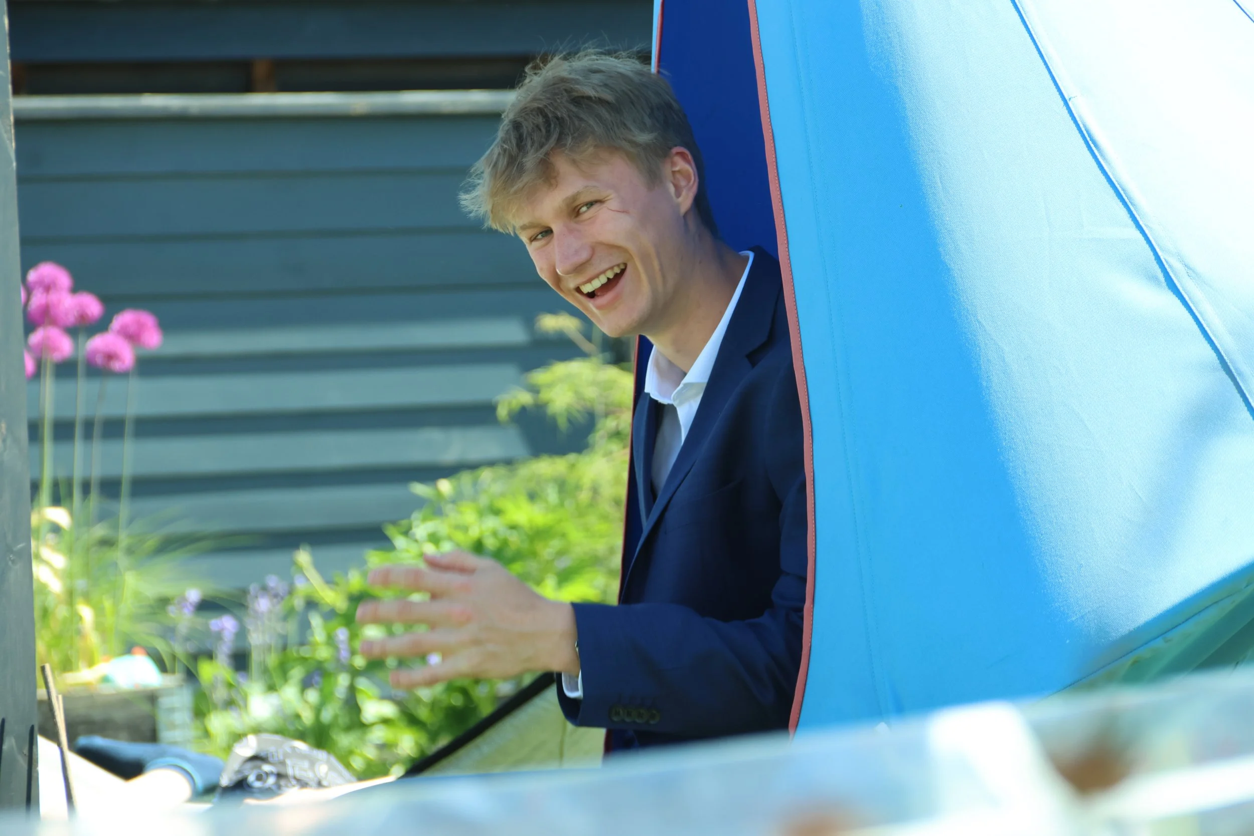 Man in a dark suit smiling and waving from a blue play tent in a garden with pink and purple flowers.
