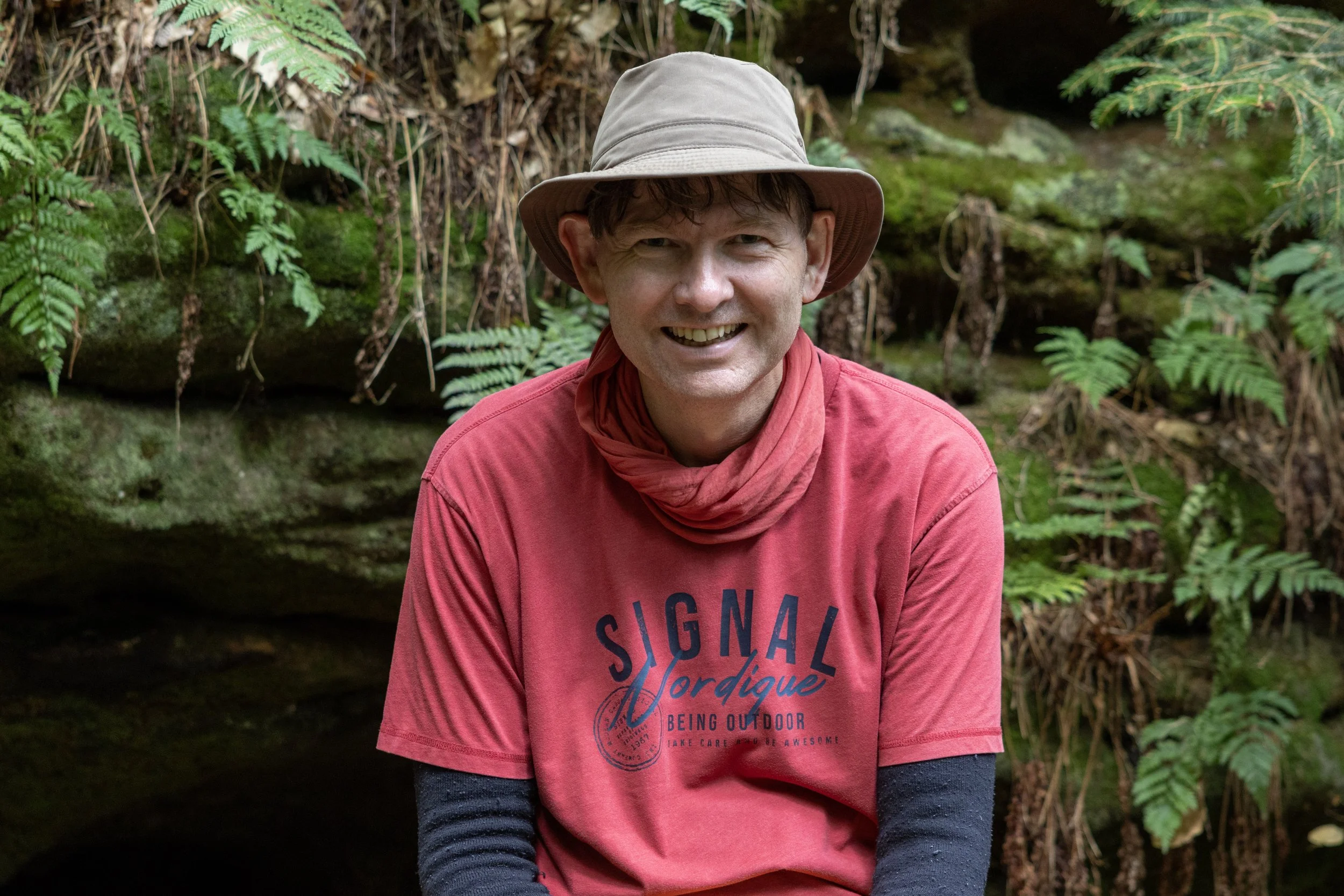 A man wearing a beige wide-brimmed hat, red t-shirt with dark sleeves underneath, and a red scarf around his neck, smiling while sitting outdoors in front of a moss-covered rock and green ferns.