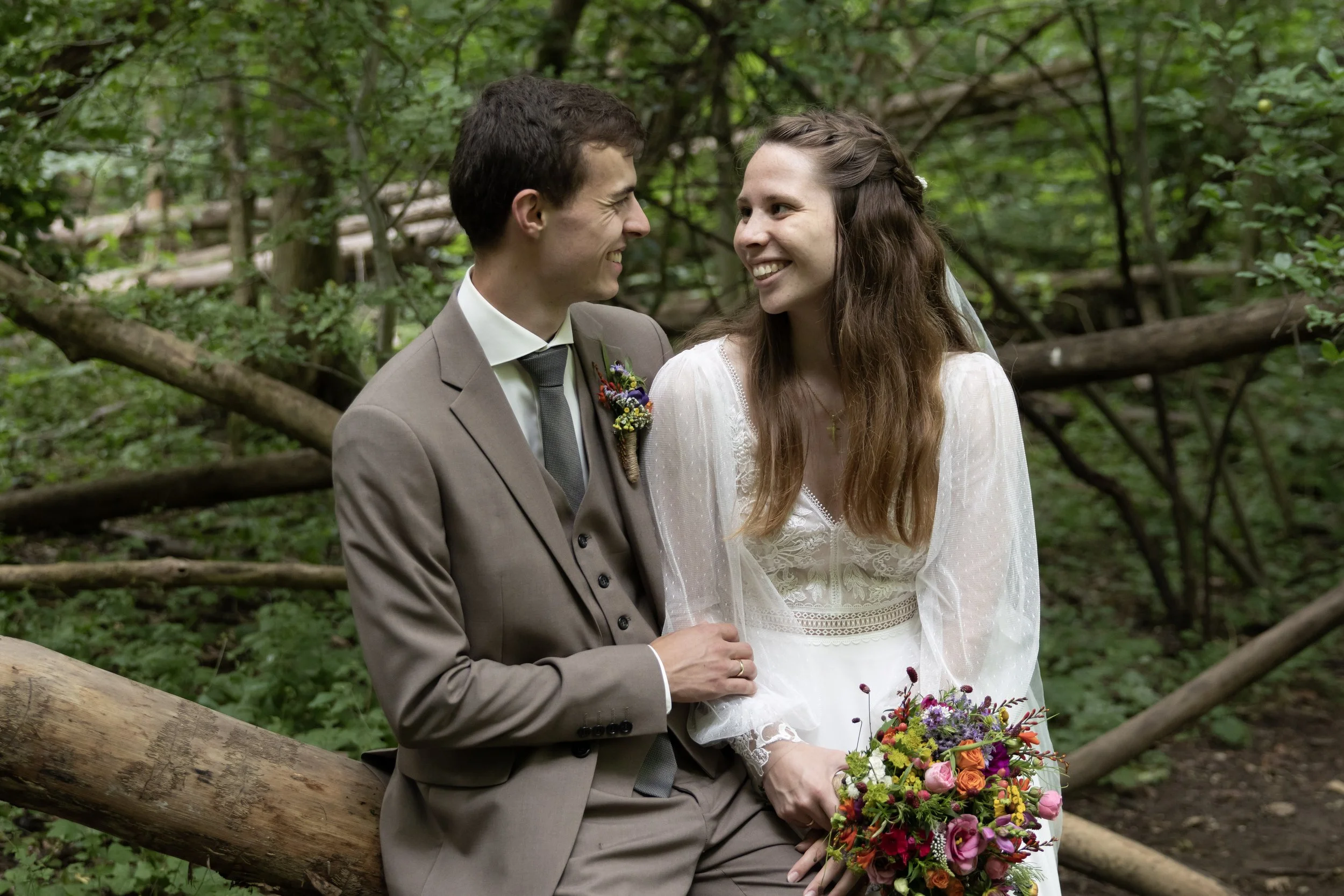 A couple dressed in wedding attire sitting on a fallen log in a lush green forest, smiling at each other. The woman holds a colorful bouquet of flowers.