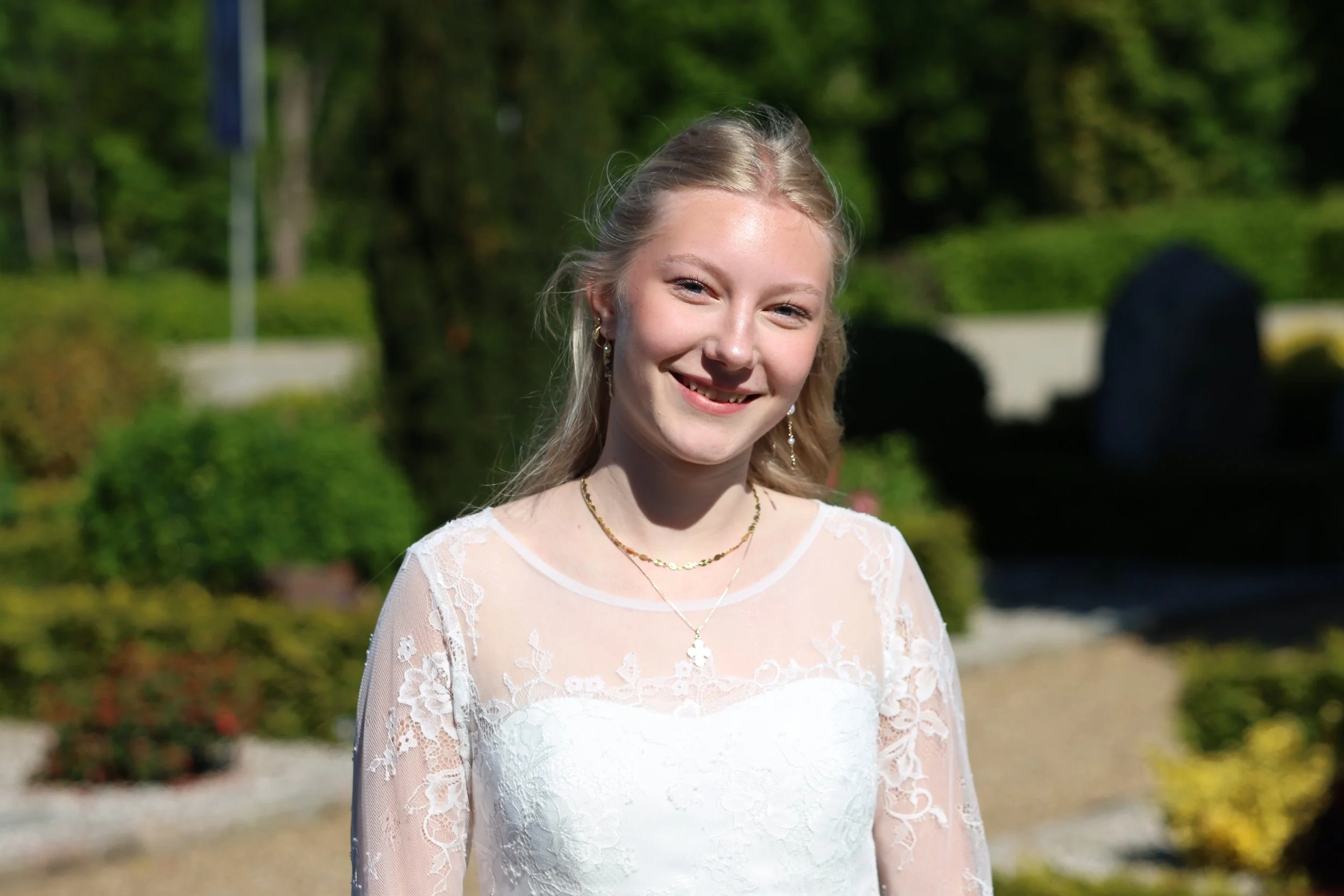 A young woman with long blonde hair wearing a white lace dress, jewelry, and earrings outdoors on a sunny day, smiling at the camera.