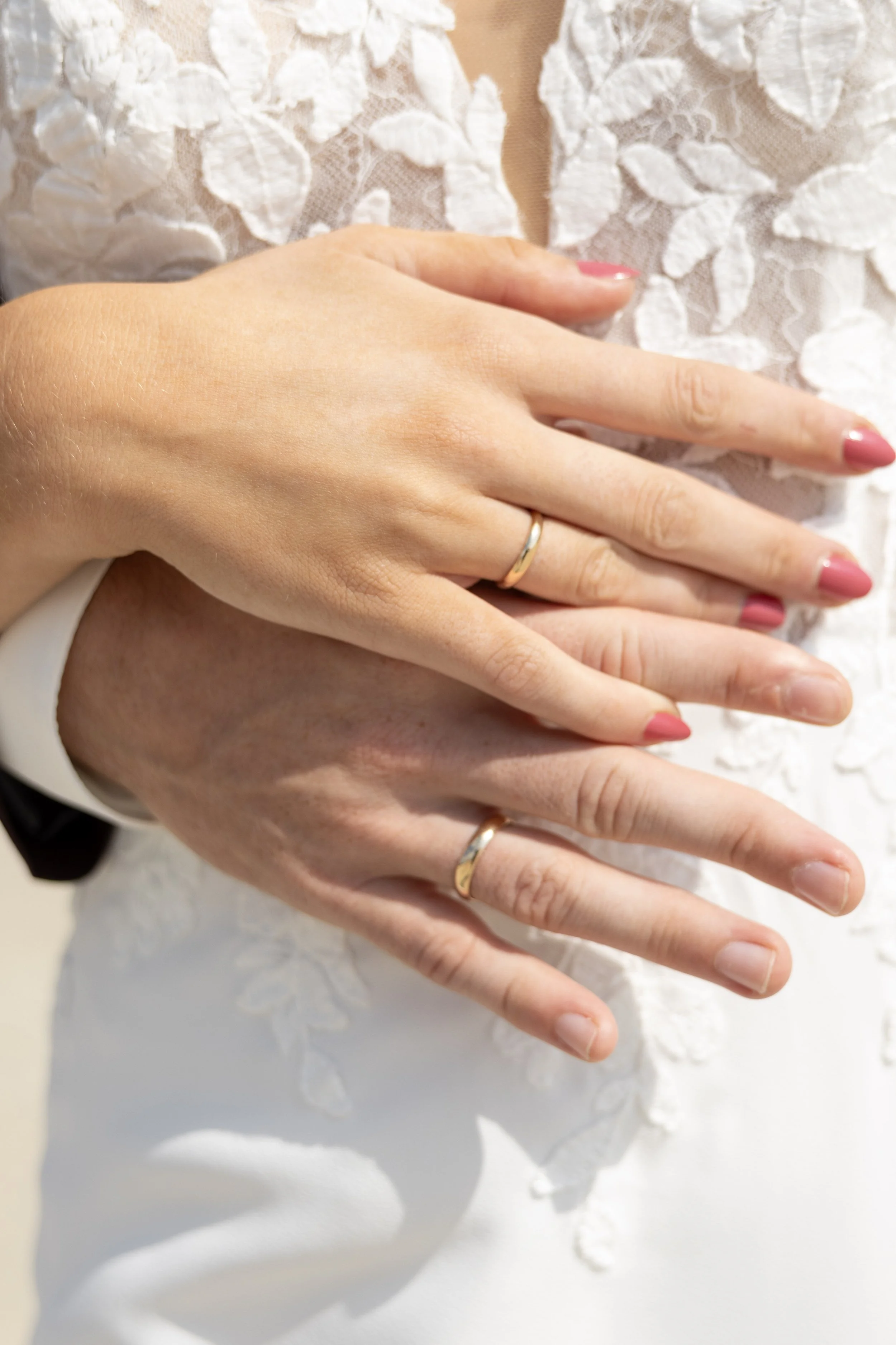 Close-up of a newlywed couple's hands with wedding rings, hands overlapping, against a white lace wedding dress.