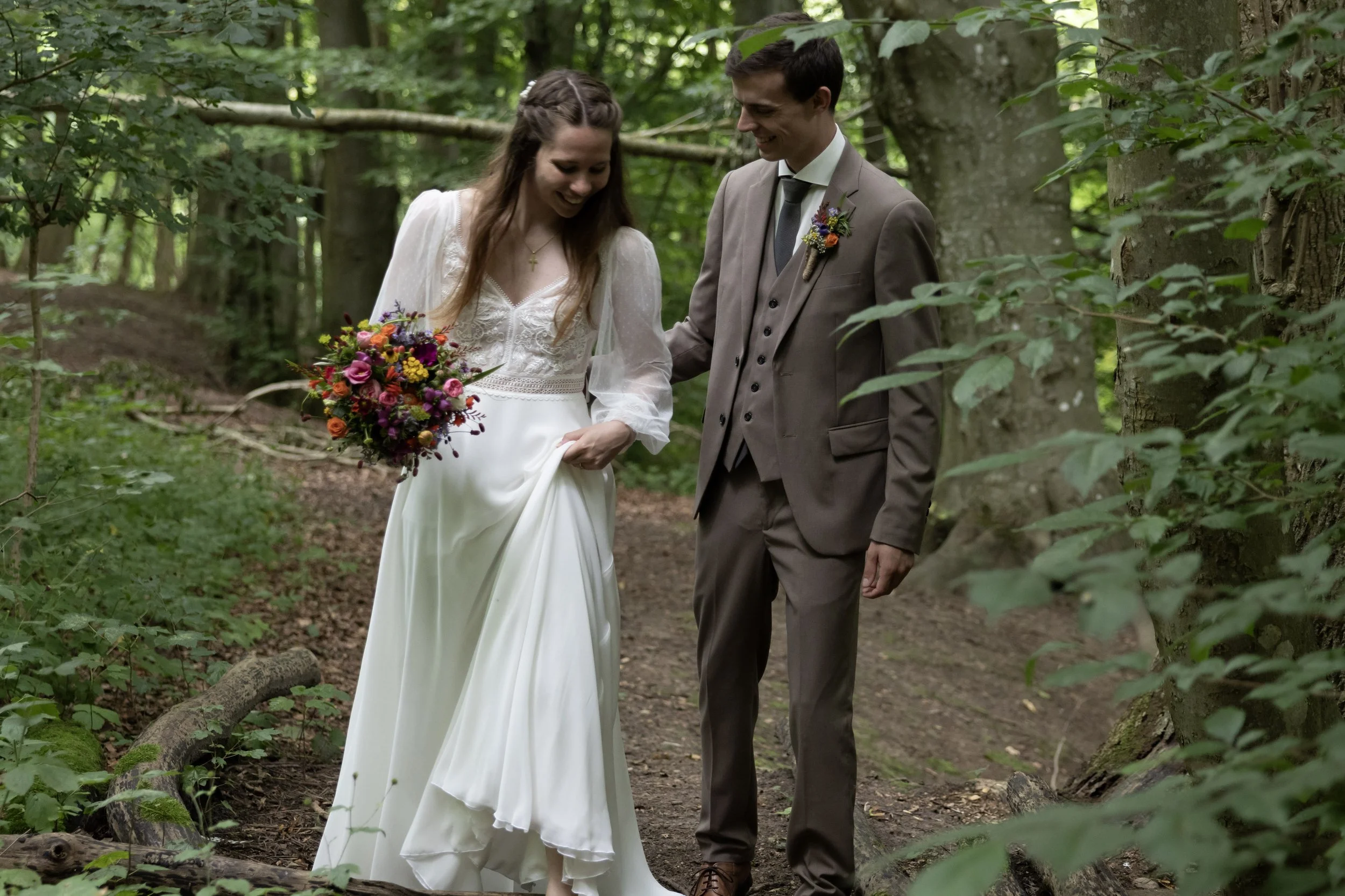 A bride and groom walking through a wooded forest, smiling and holding hands, with the bride holding a colorful bouquet of flowers.