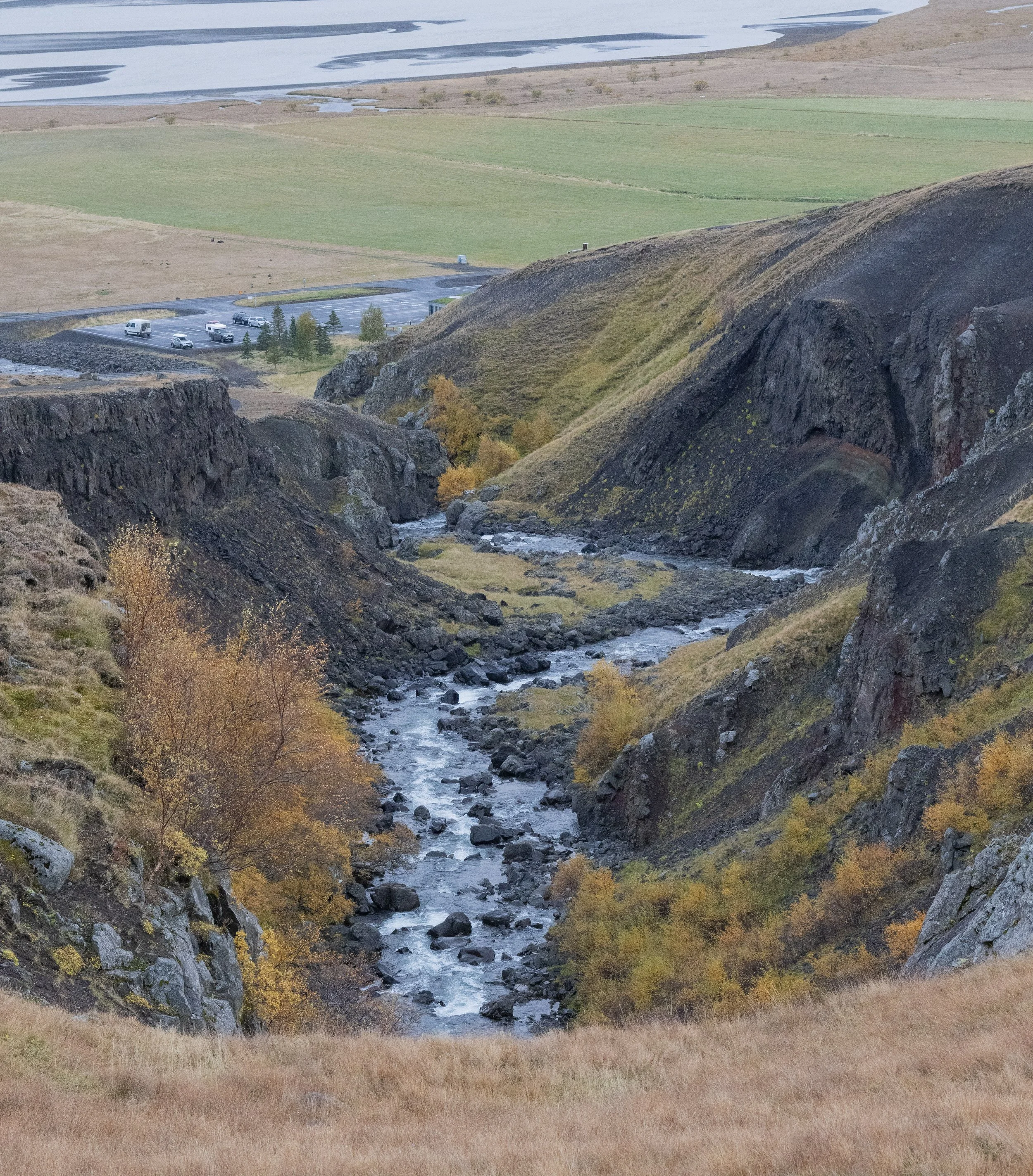 A narrow river flows through a rocky canyon with steep black and green hillside slopes on either side. In the background, there are green fields and a parking lot with several cars, separated from the canyon by a small road.