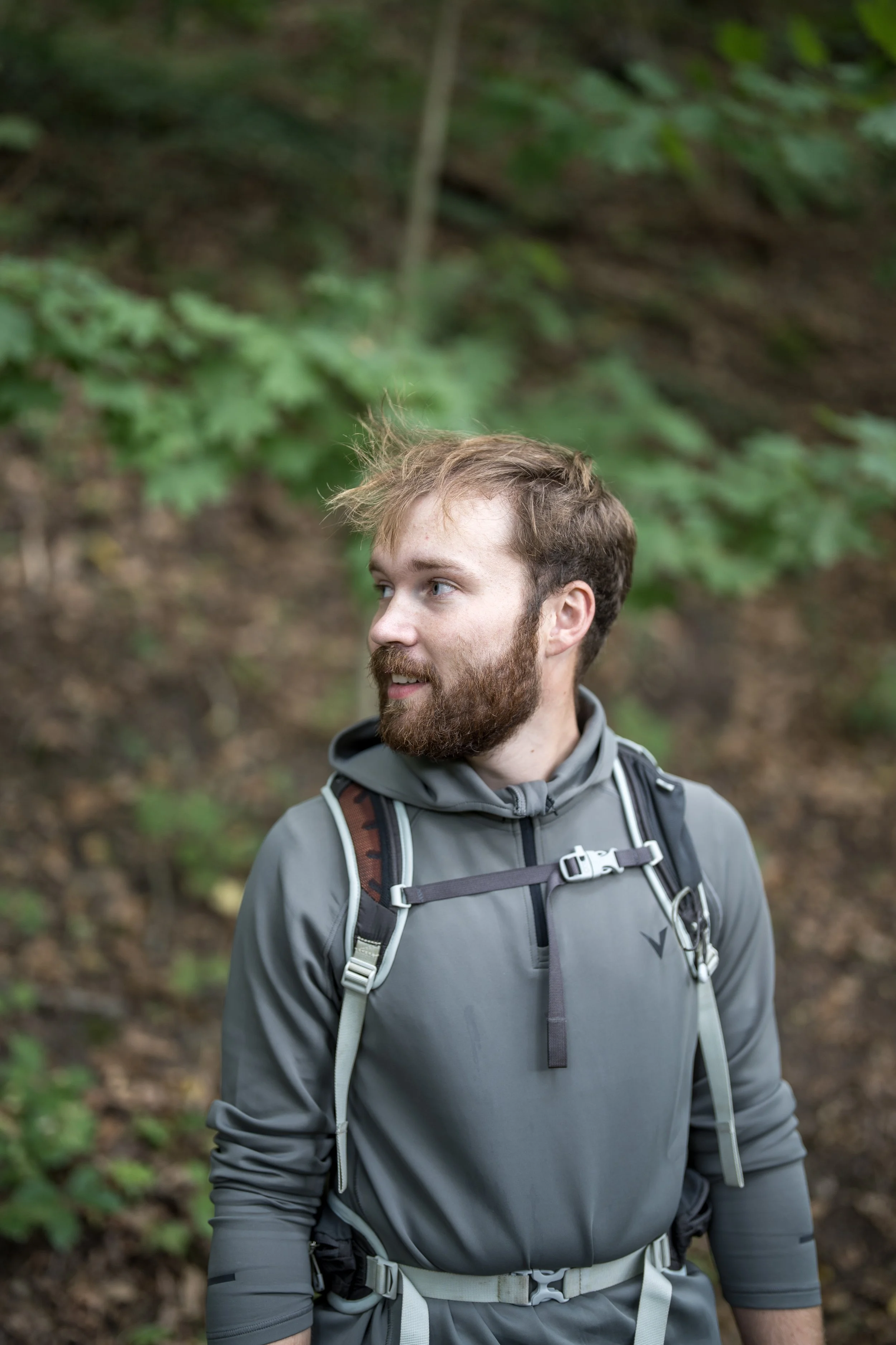 A man with a beard, wearing a gray jacket and backpack, standing outdoors in a wooded area.