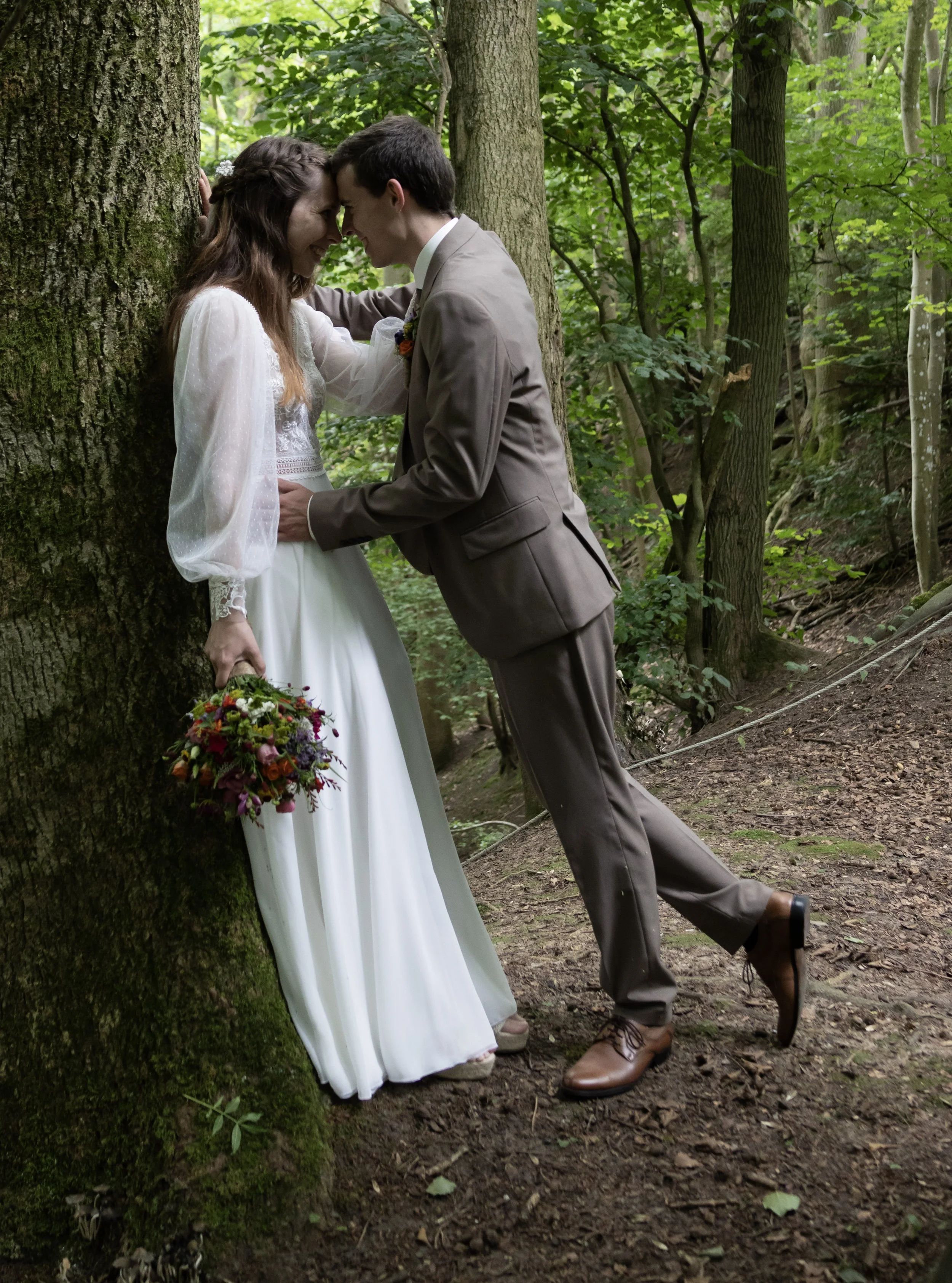 A bride and groom in wedding attire leaning against a tree in a forest, with foreheads touching and eyes closed, holding a bouquet of colorful flowers.