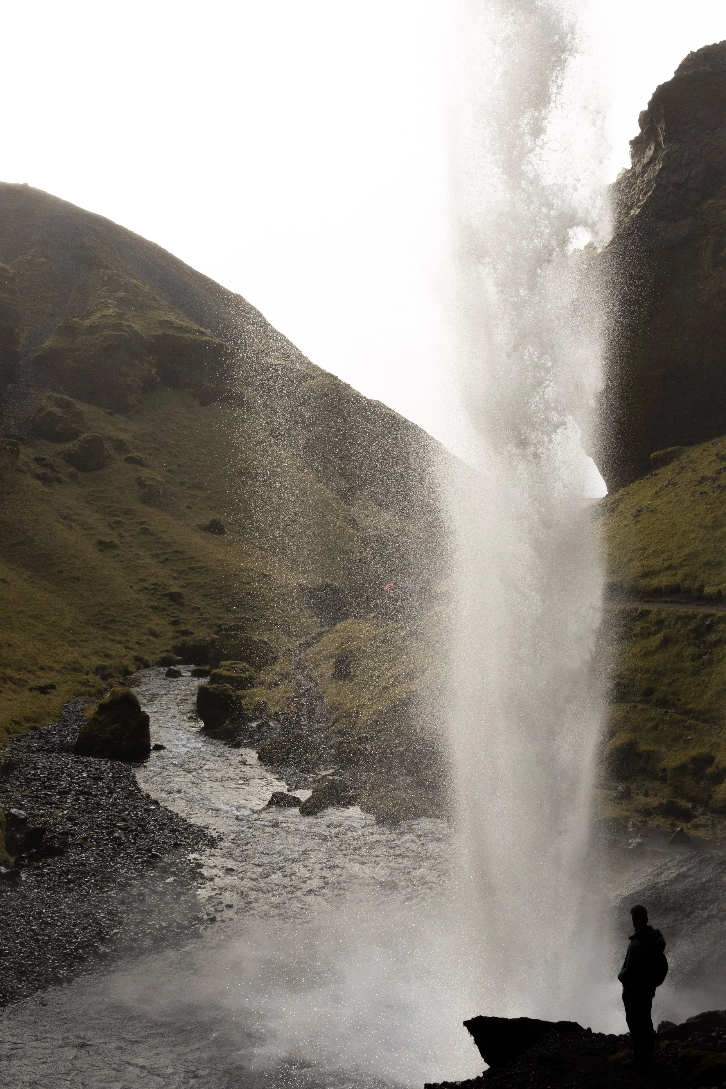A person standing on rocks near a large waterfall in a valley with green moss-covered hills on either side.