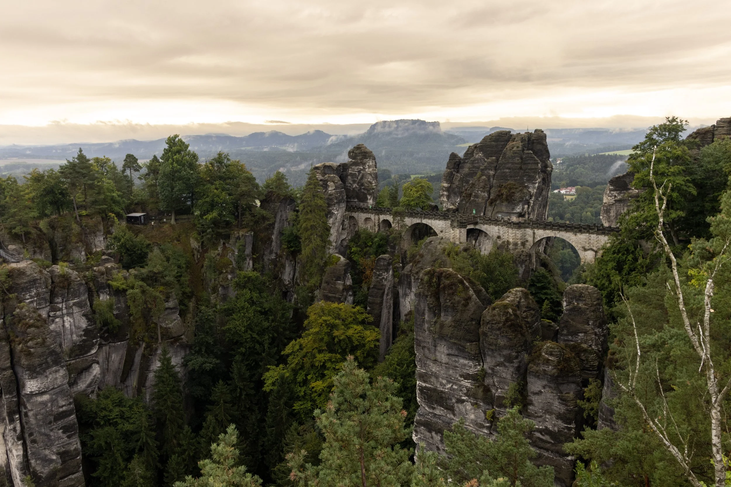 Scenic view of a stone bridge spanning across tall rock formations in a lush green forest with mountains and cloudy sky in the background.