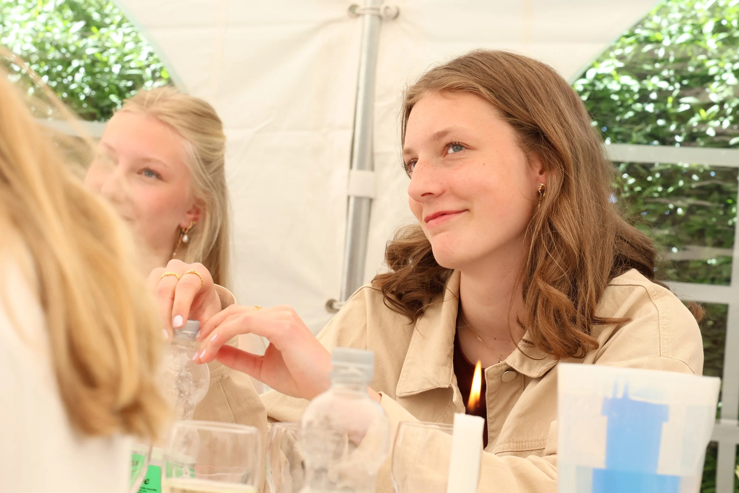 Two young women with light brown hair and light skin sitting at a table outdoors under a tent, smiling and engaging in an activity involving bottles and candles, with greenery in the background.