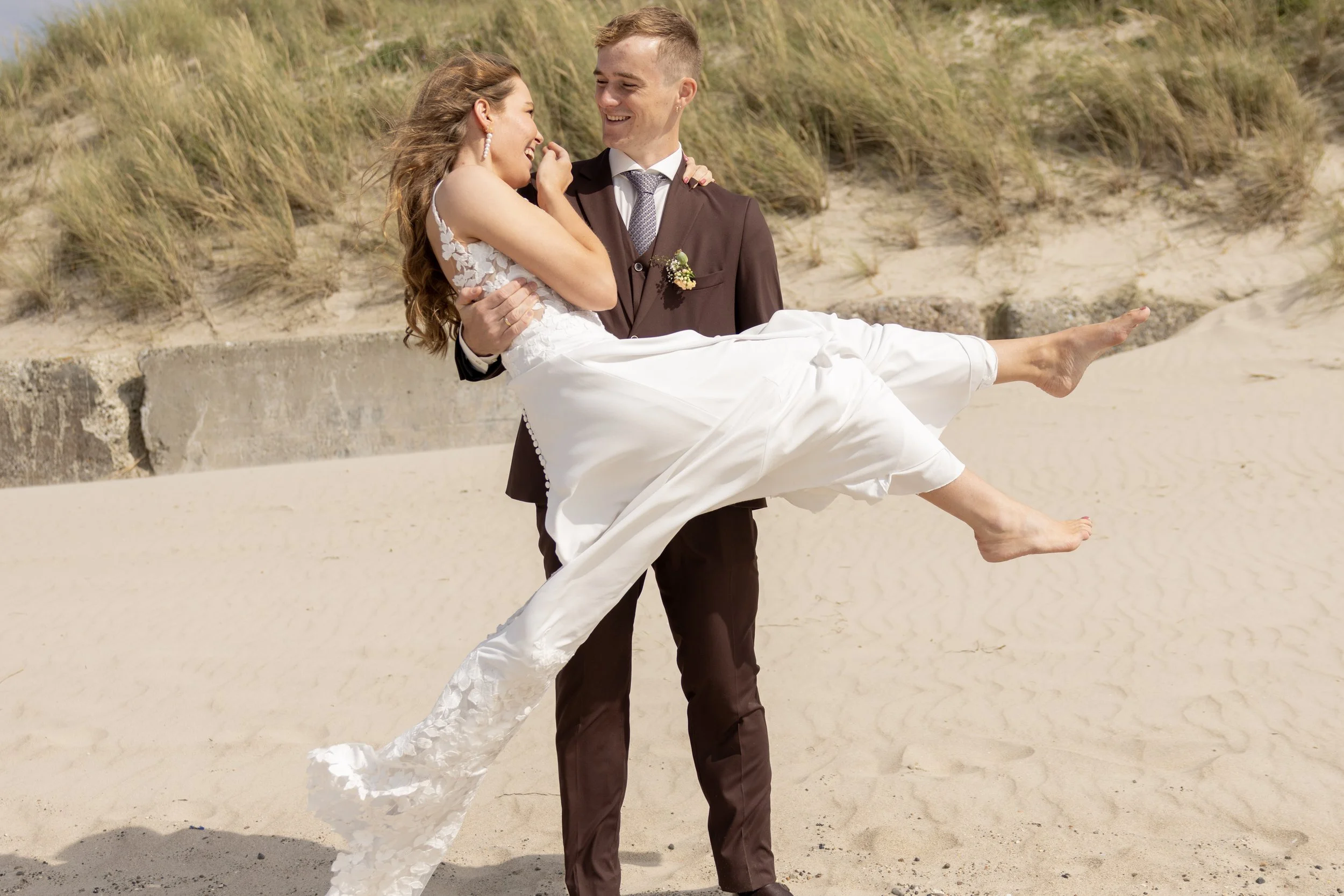 A groom in a brown suit holding a bride in a white wedding dress on a sandy beach with grassy dunes in the background.