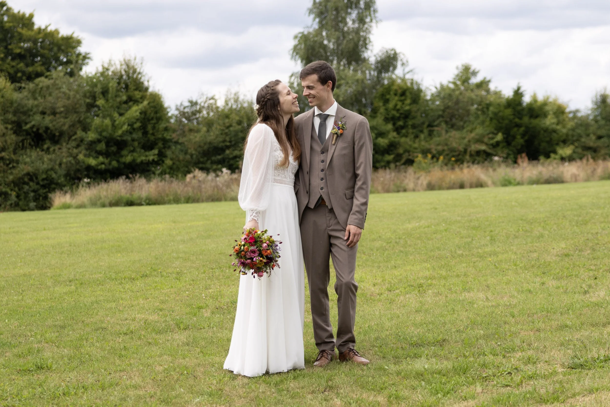 A bride and groom standing on a grassy field, smiling and looking at each other. The bride holds a colorful bouquet, and the groom wears a boutonniere. They are outdoors with trees and a cloudy sky in the background.