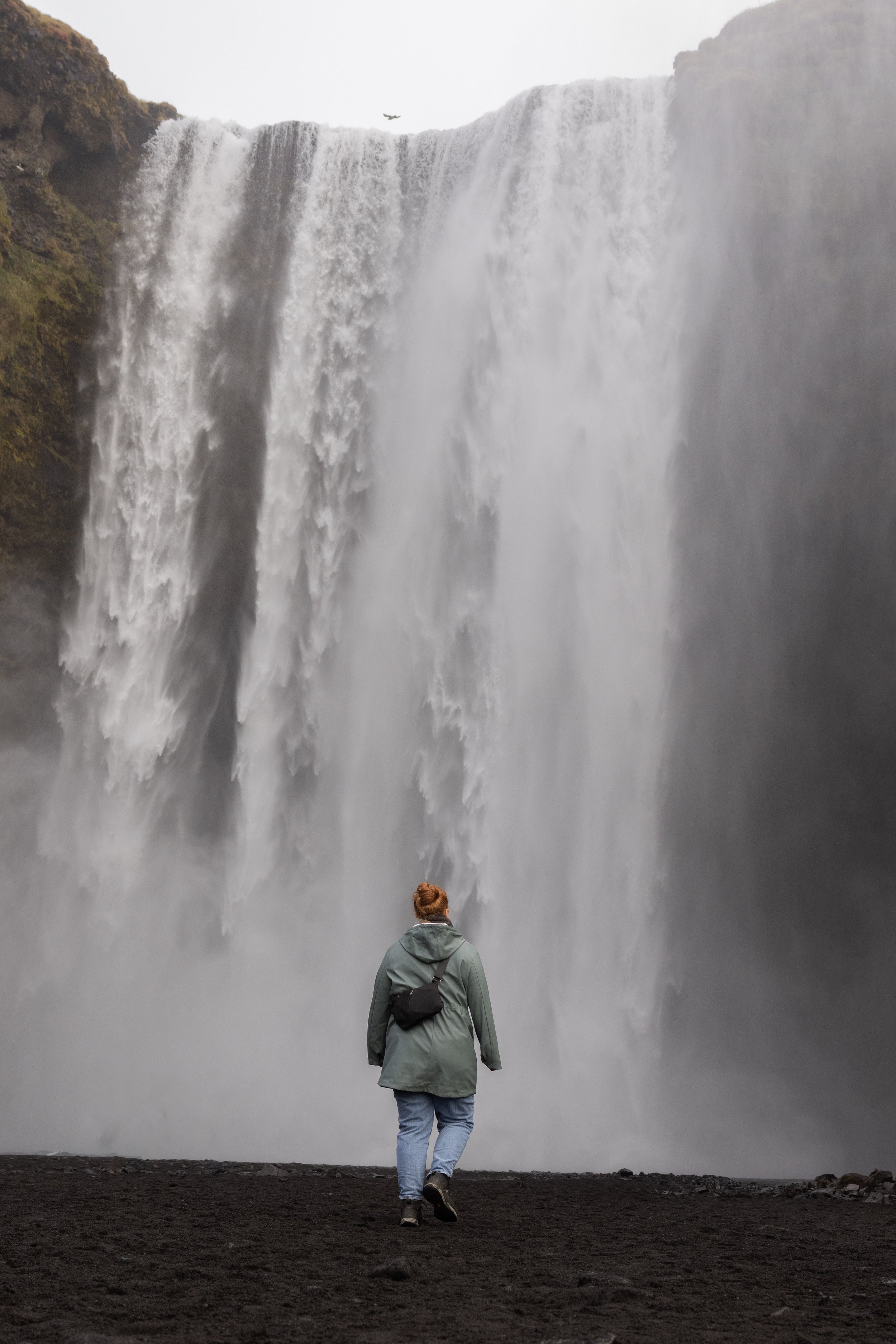 A person with red hair wearing a green jacket, jeans, and a black backpack walking toward a large waterfall, with mist and water spray visible.