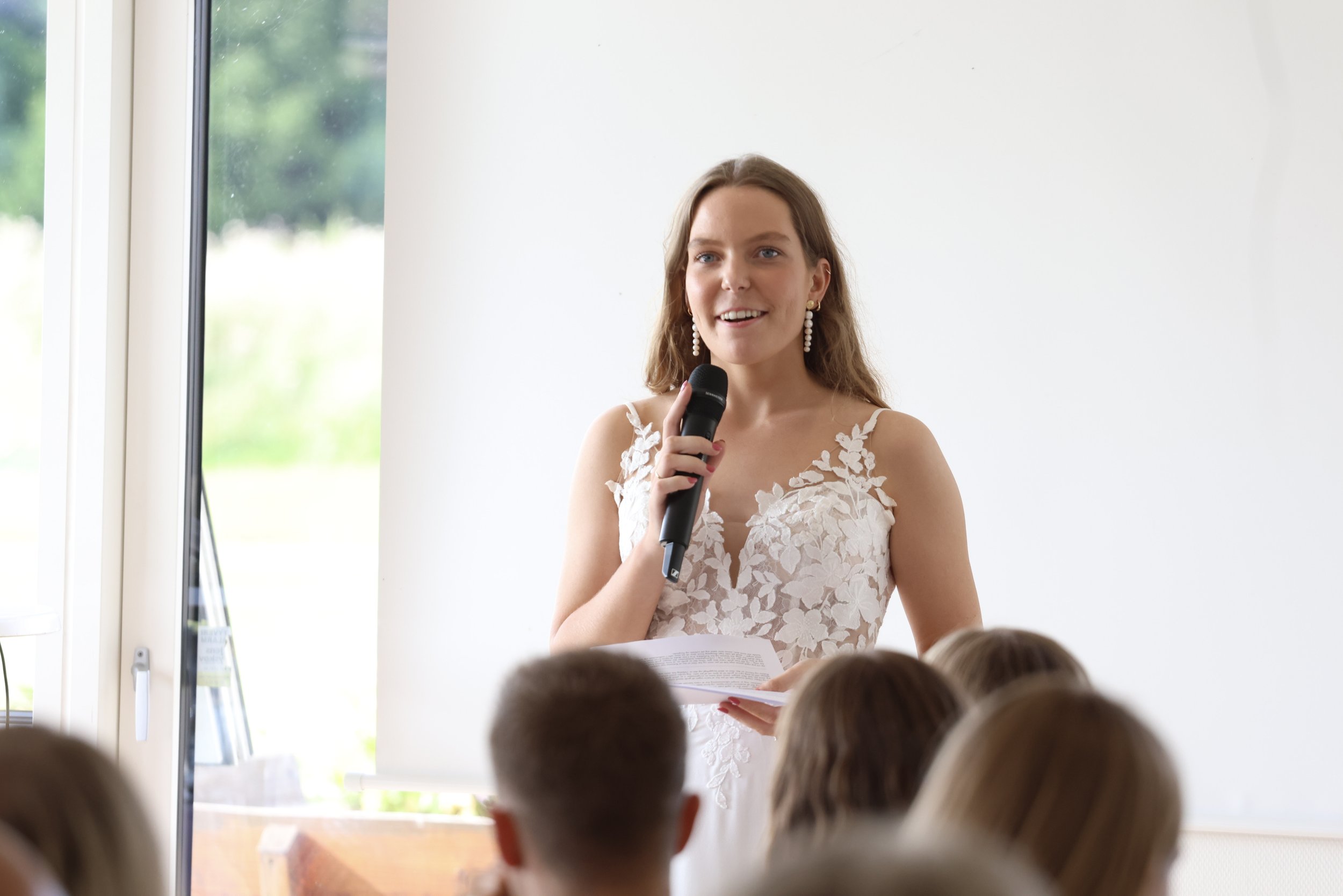 A young woman in a white lace dress holding a microphone and a script, speaking to an audience in a well-lit room with large windows.