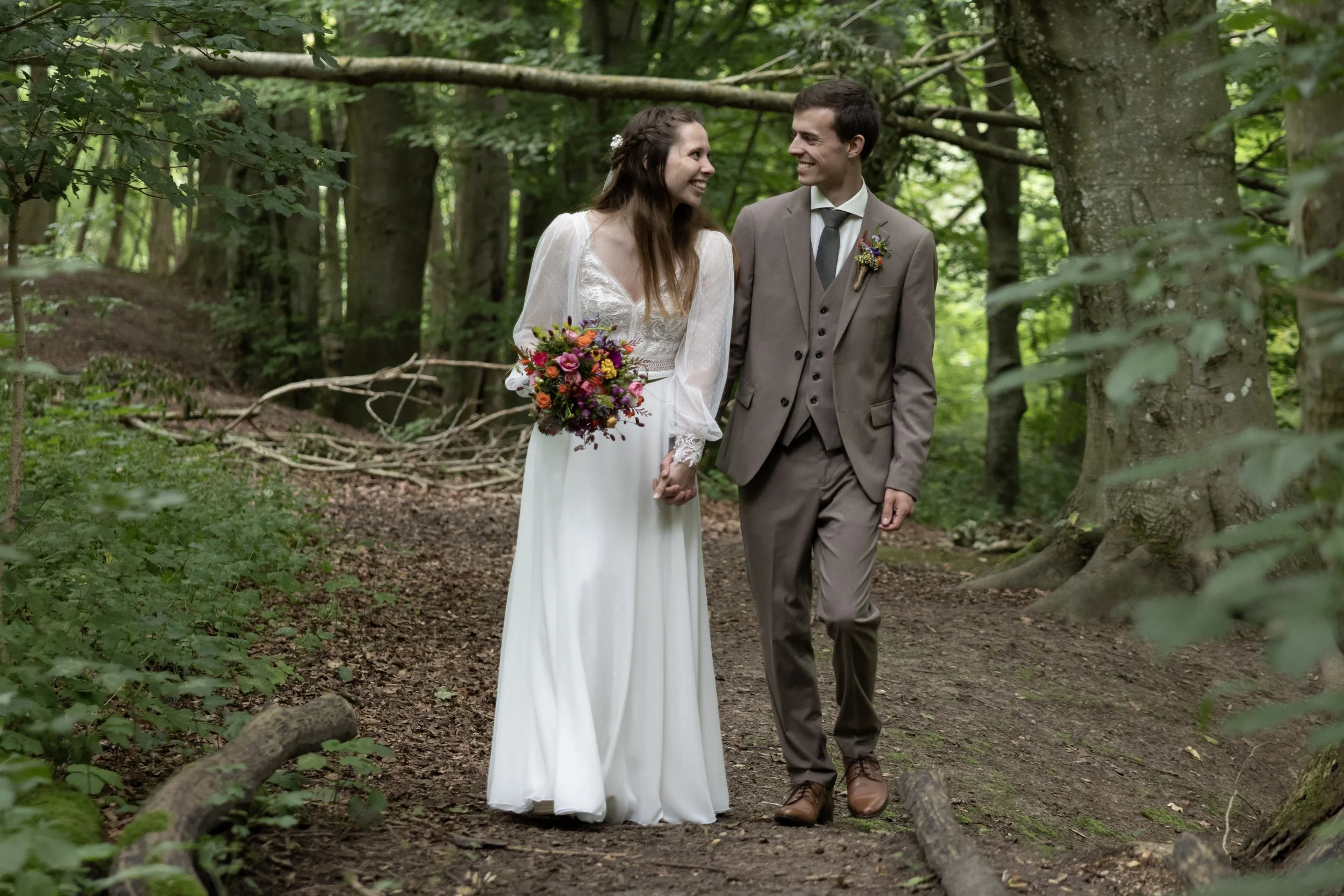 A bride and groom walk hand in hand through a lush green forest, smiling at each other. The bride wears a white dress and holds a colorful bouquet; the groom wears a brown suit and tie.
