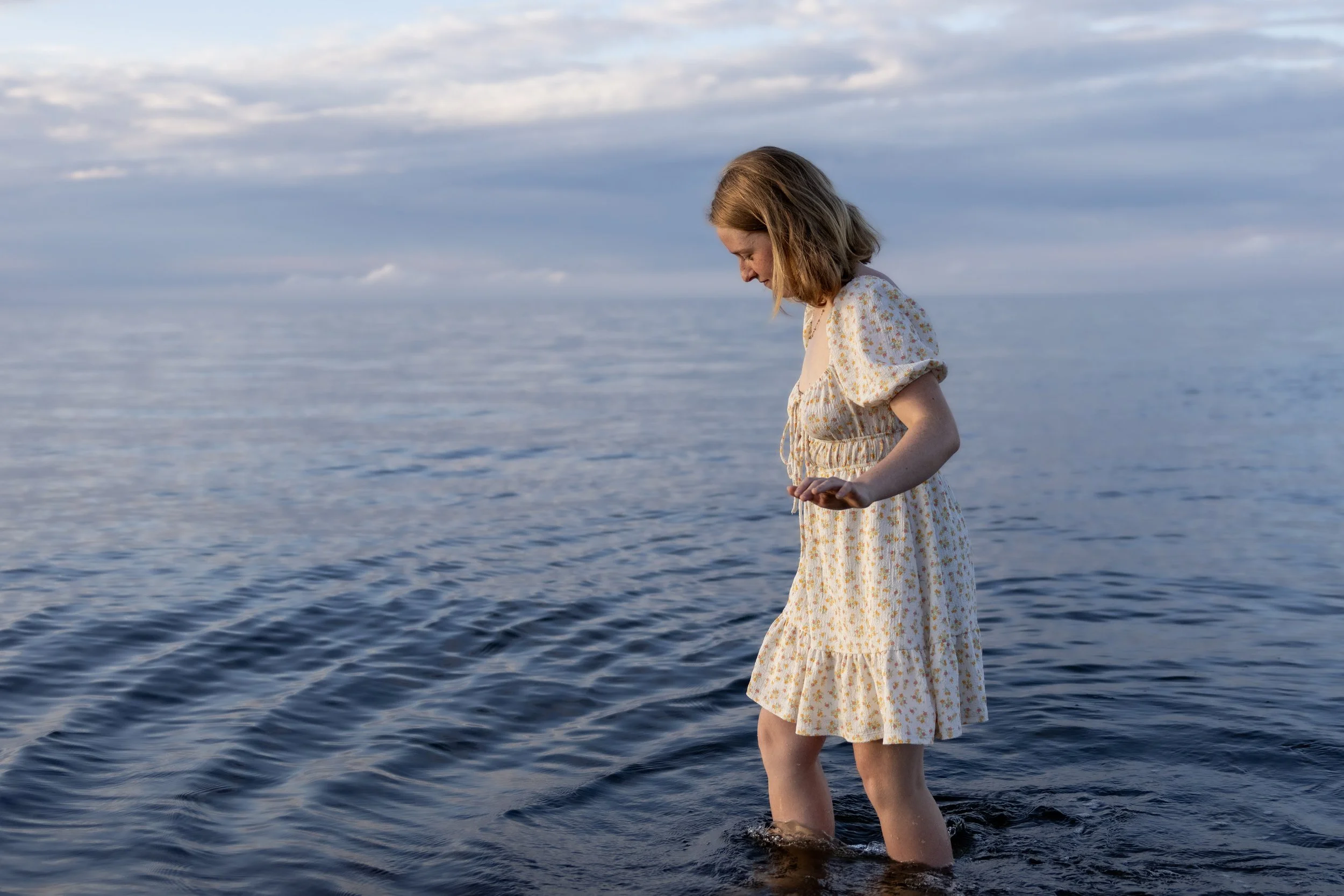 A young girl in a floral dress standing in the shallow water of a calm lake or ocean, looking down at the water, with mostly cloudy sky in the background.