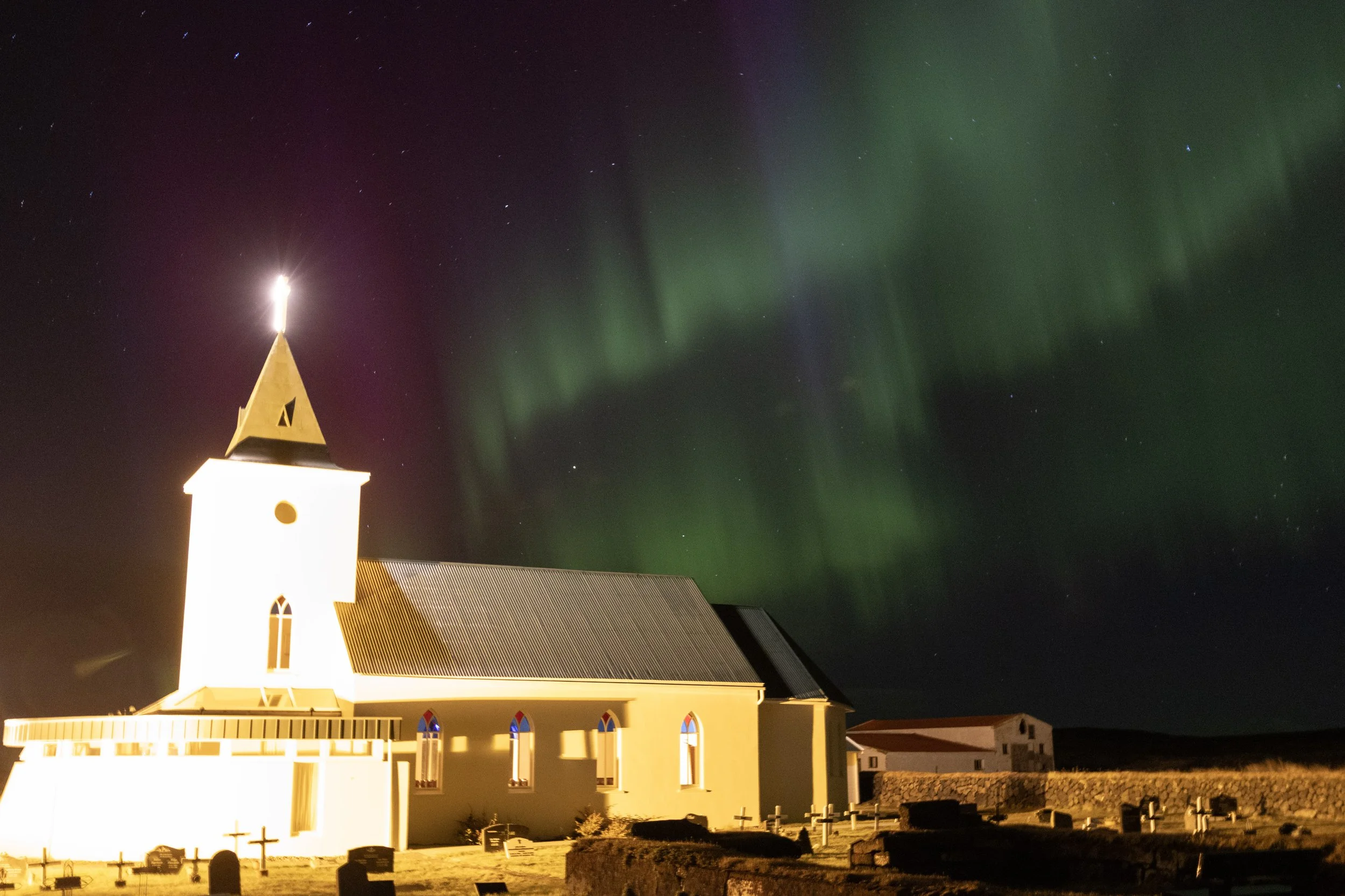 Nighttime scene of a white church with a tall steeple, illuminated from below, under a sky displaying green northern lights (aurora borealis).