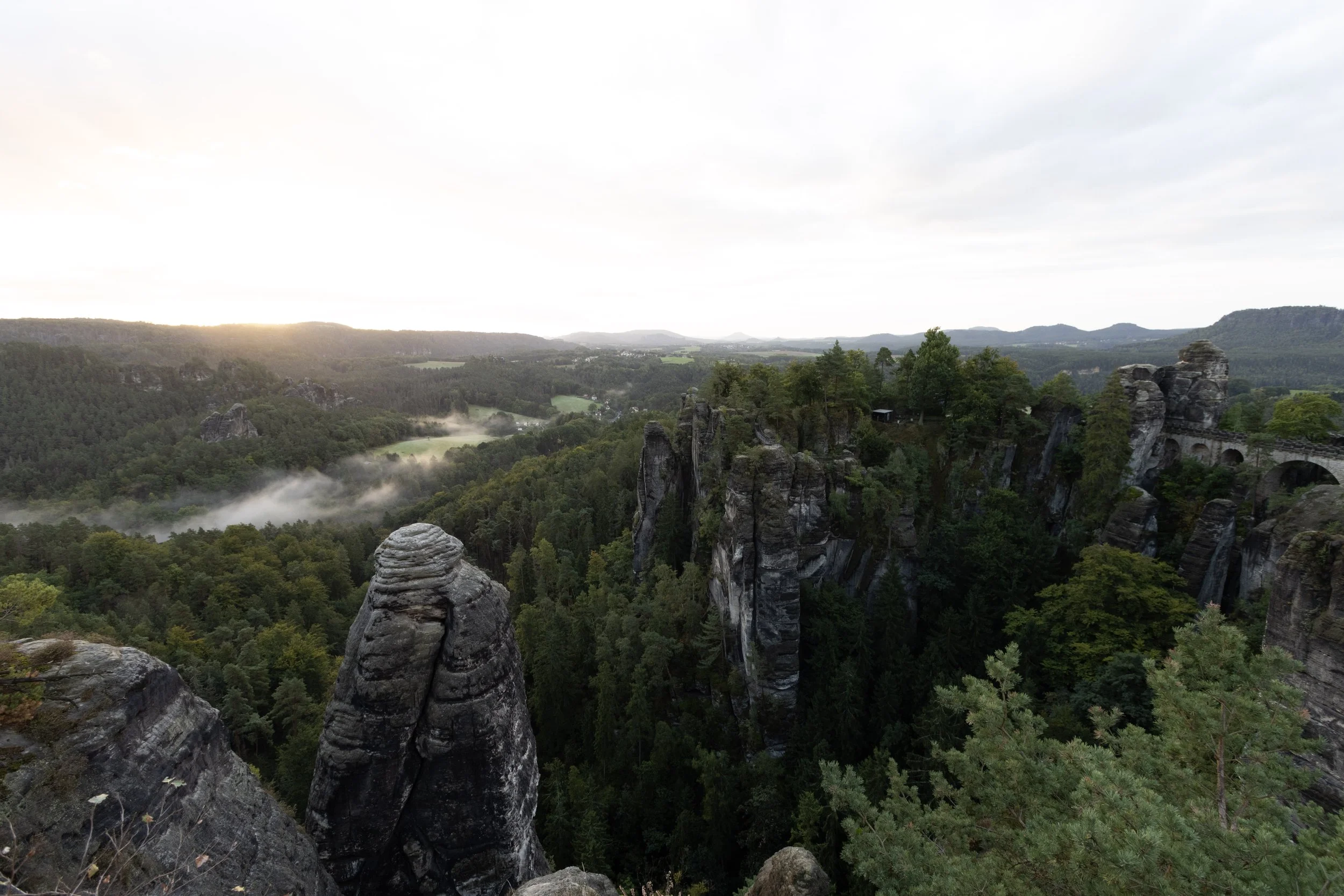Scenic view of rock formations and a forested valley during sunrise or sunset with mist and rolling hills in the distance.