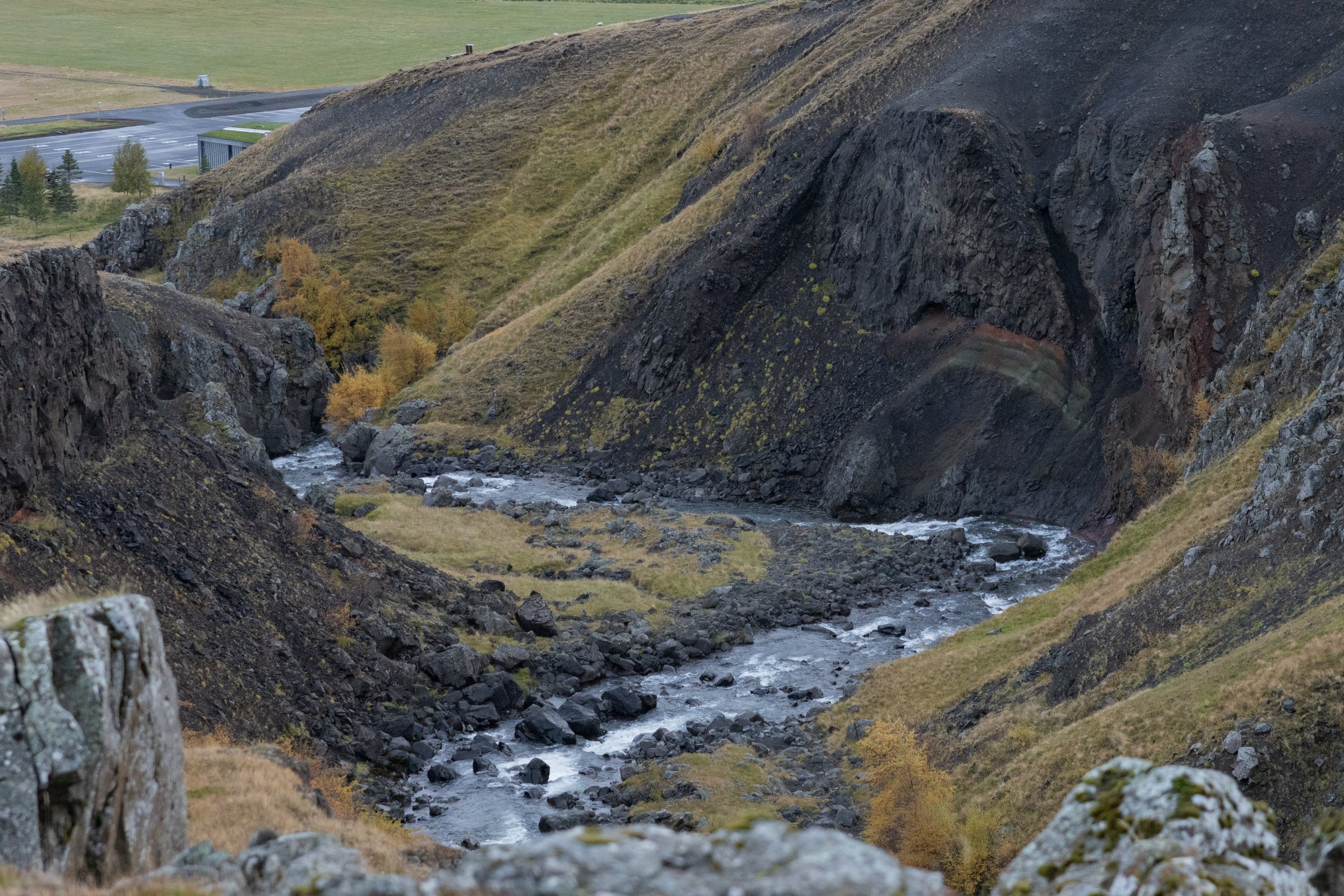 A narrow river flowing through a rocky, steep canyon with grassy slopes and sparse trees, with a distant parking lot and grassy field in the background.