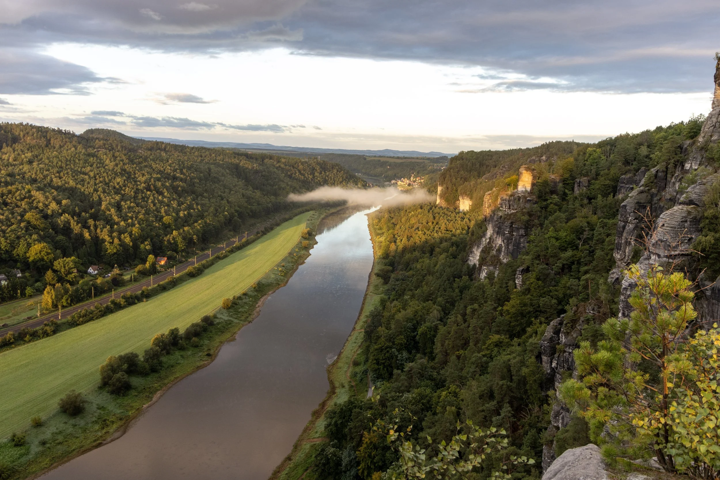 A river flanked by green fields and dense forests on both sides, with stone cliffs on the right, in a hilly landscape under a partly cloudy sky.