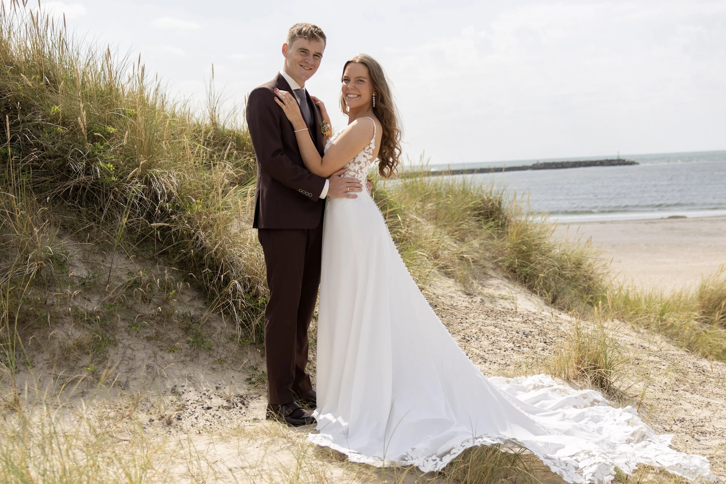 A bride and groom standing on a sandy beach with grass-covered dunes, smiling, with the ocean and sky in the background.