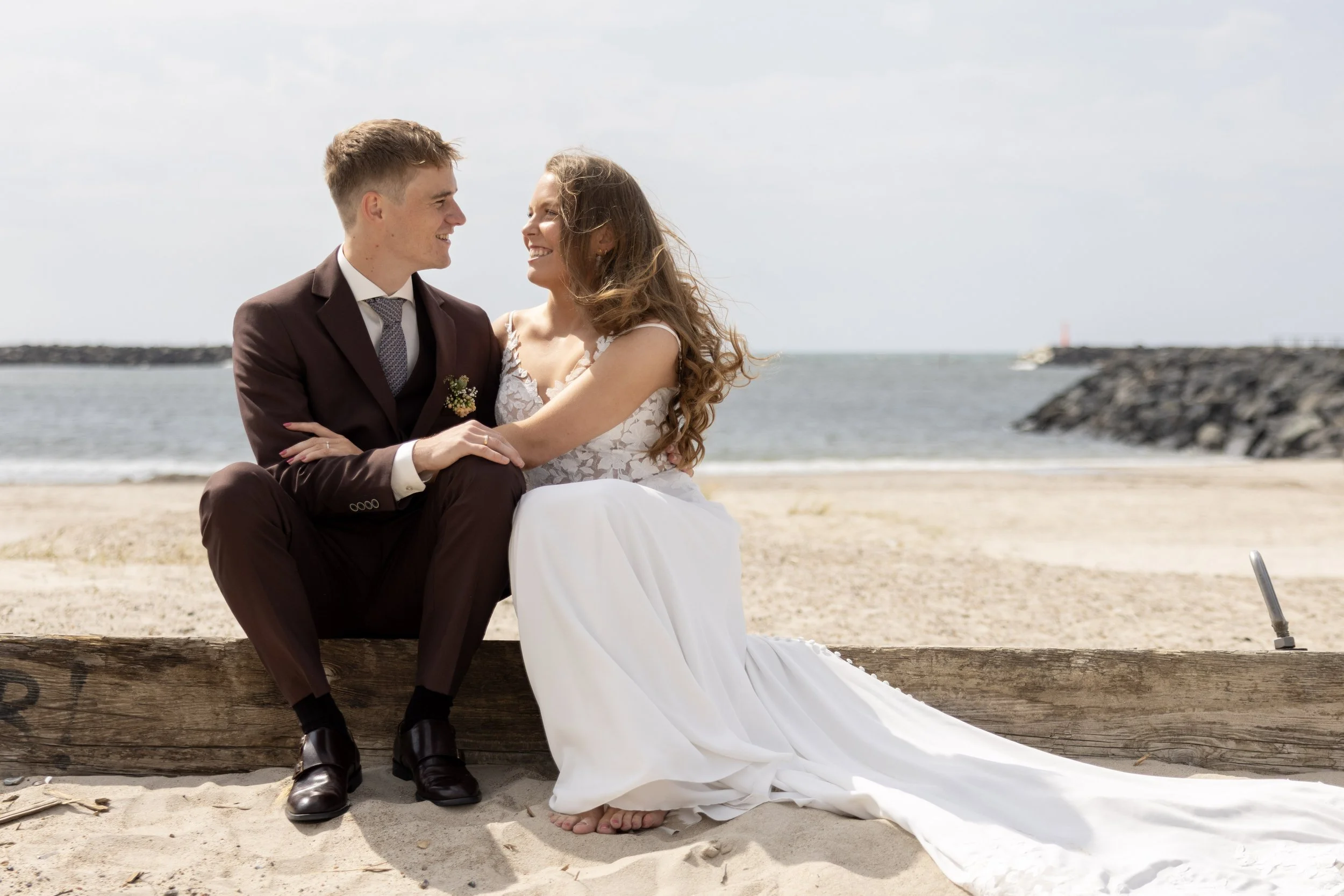 A newlywed couple sits on a wooden beam on a sandy beach, smiling and looking into each other's eyes with the ocean and sky in the background.