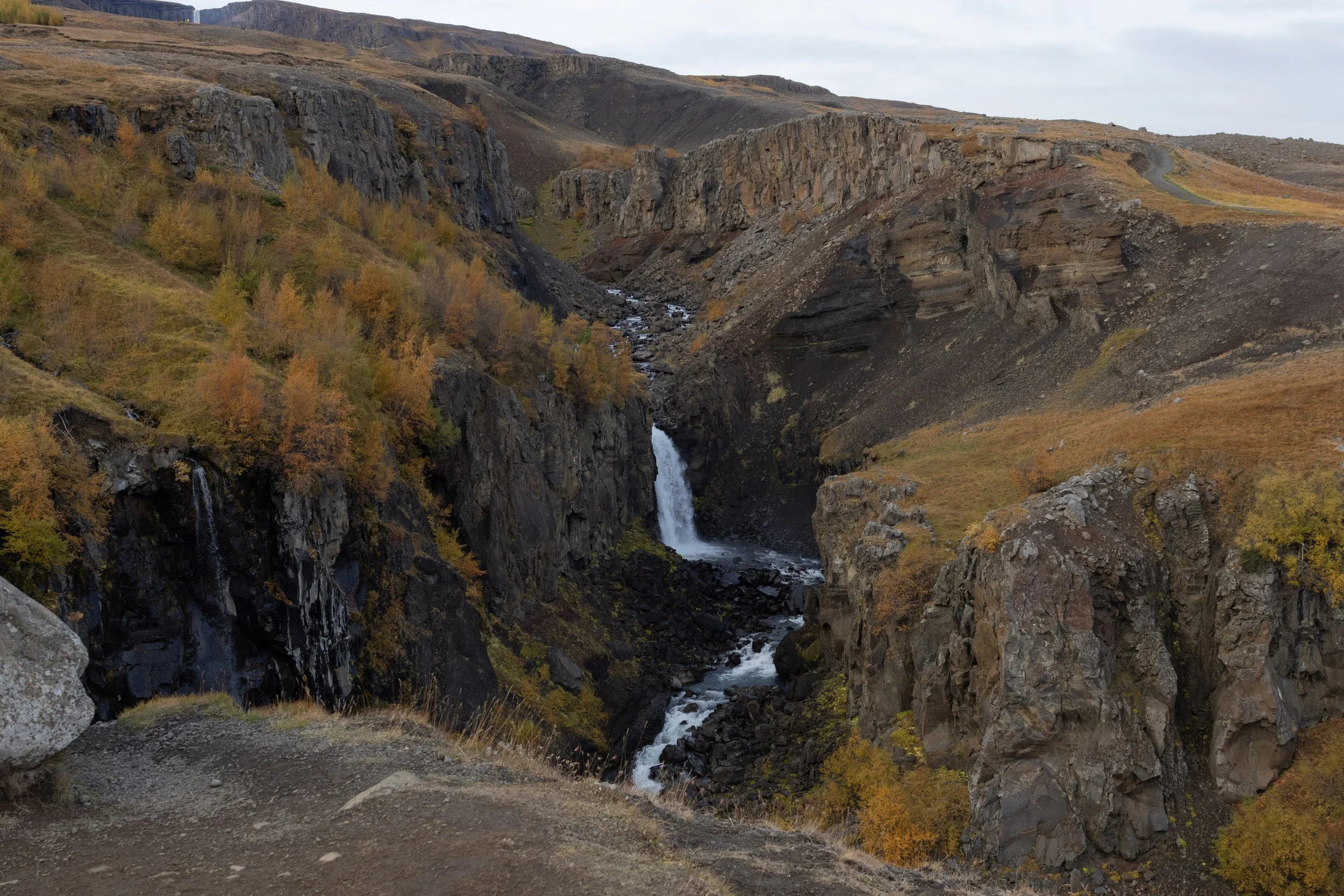 A waterfall cascading down rocky cliffs amidst autumn foliage on a cloudy day.