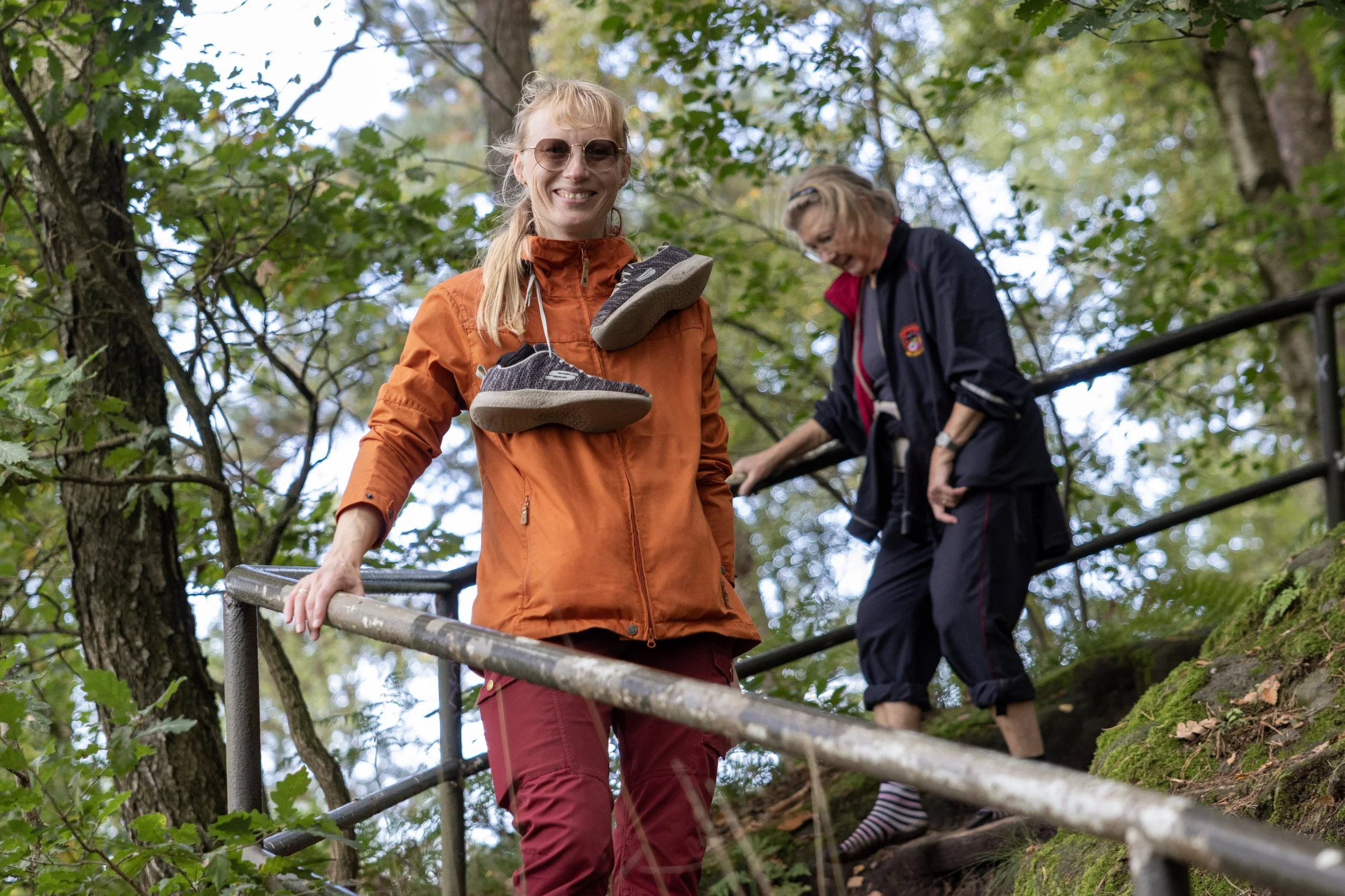 Two women descending a forest trail with a metal handrail, surrounded by trees with green leaves.