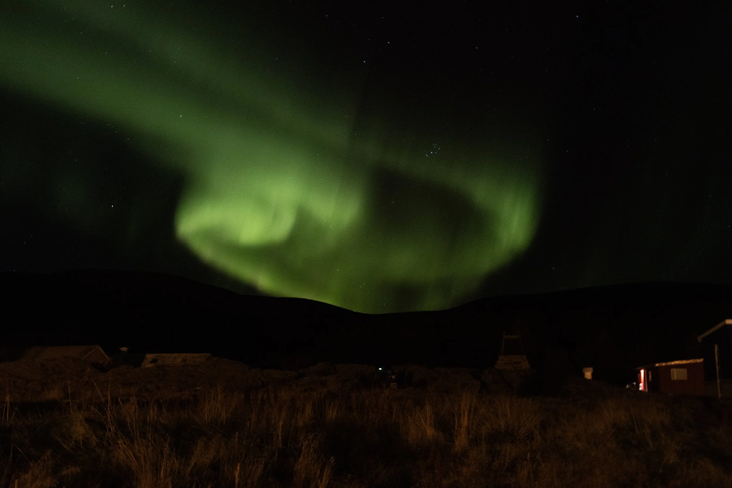 Northern lights (aurora borealis) glowing green in the night sky above a dark landscape with buildings and grass.