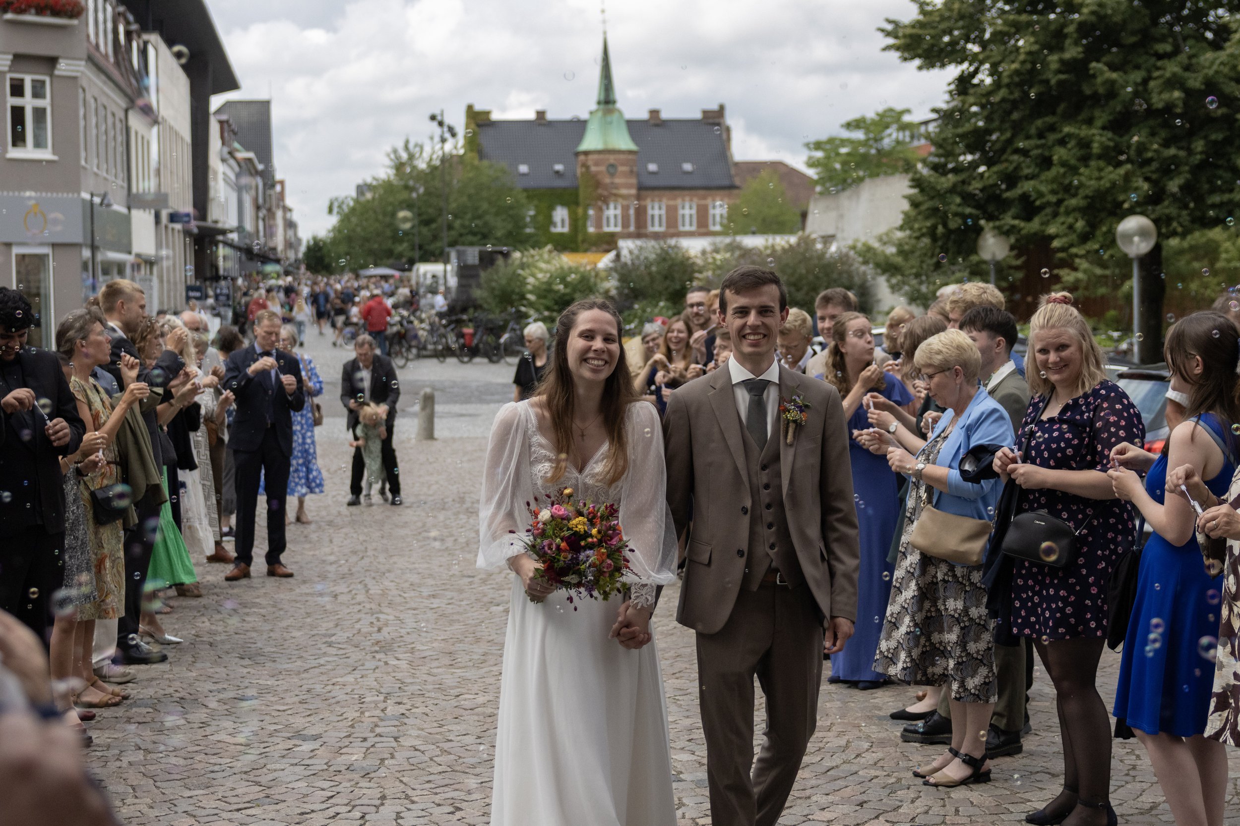 A bride and groom walking together on a cobblestone street surrounded by wedding guests, with buildings and a church tower in the background.