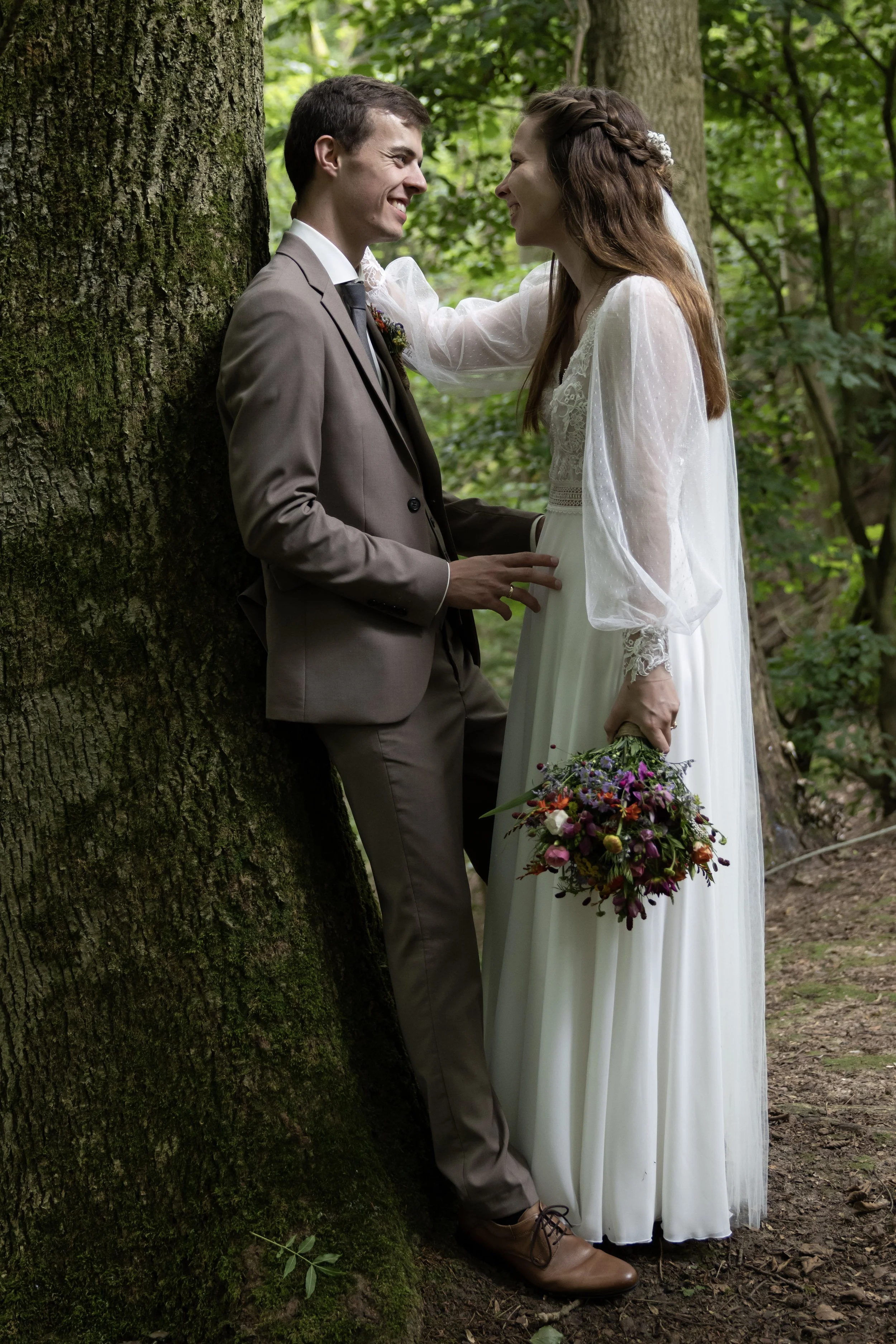 A newlywed couple standing with their foreheads and noses touching in a forest, smiling at each other. The groom is wearing a brown suit, and the bride is in a white wedding dress holding a bouquet of flowers.