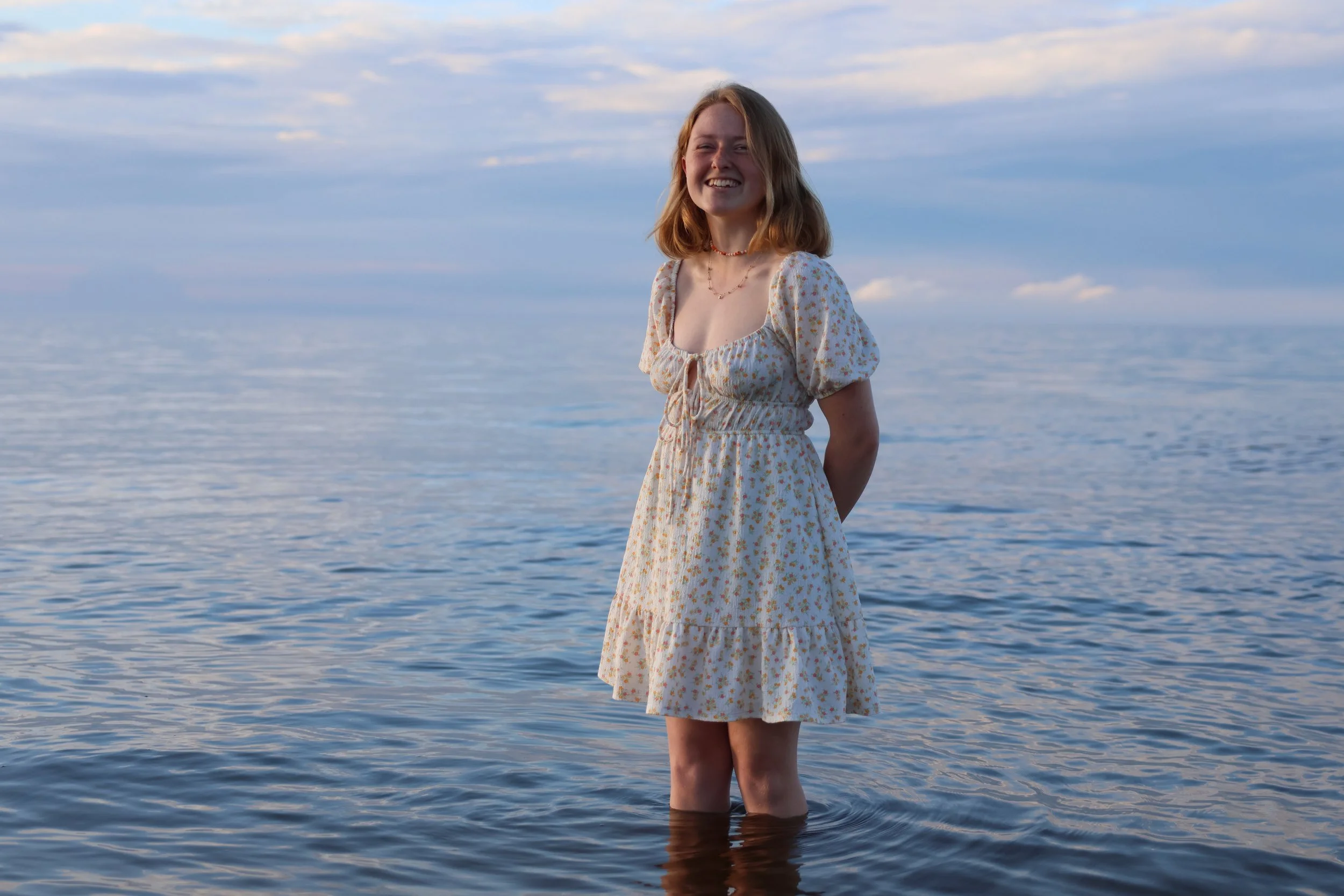 A young woman in a floral dress standing in shallow water at the beach during sunset, smiling with her eyes closed.