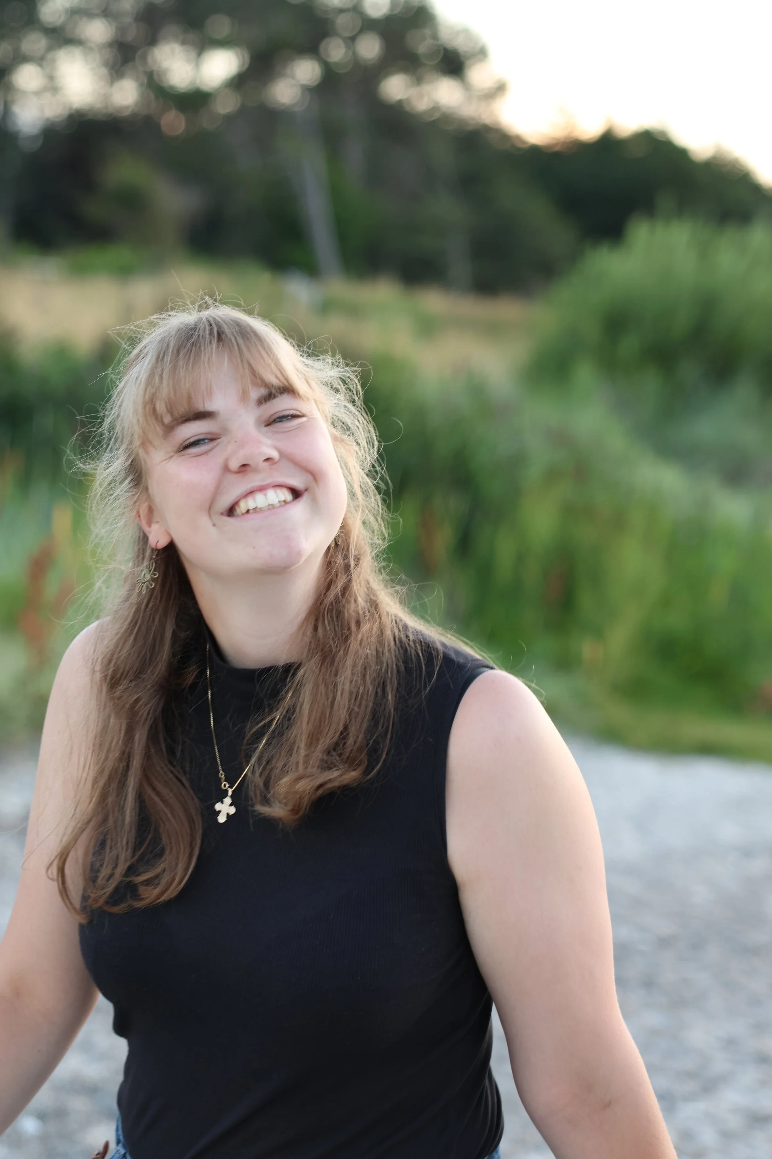 Young woman with long light brown hair and bangs smiling outdoors in a natural setting with greenery and trees in the background.