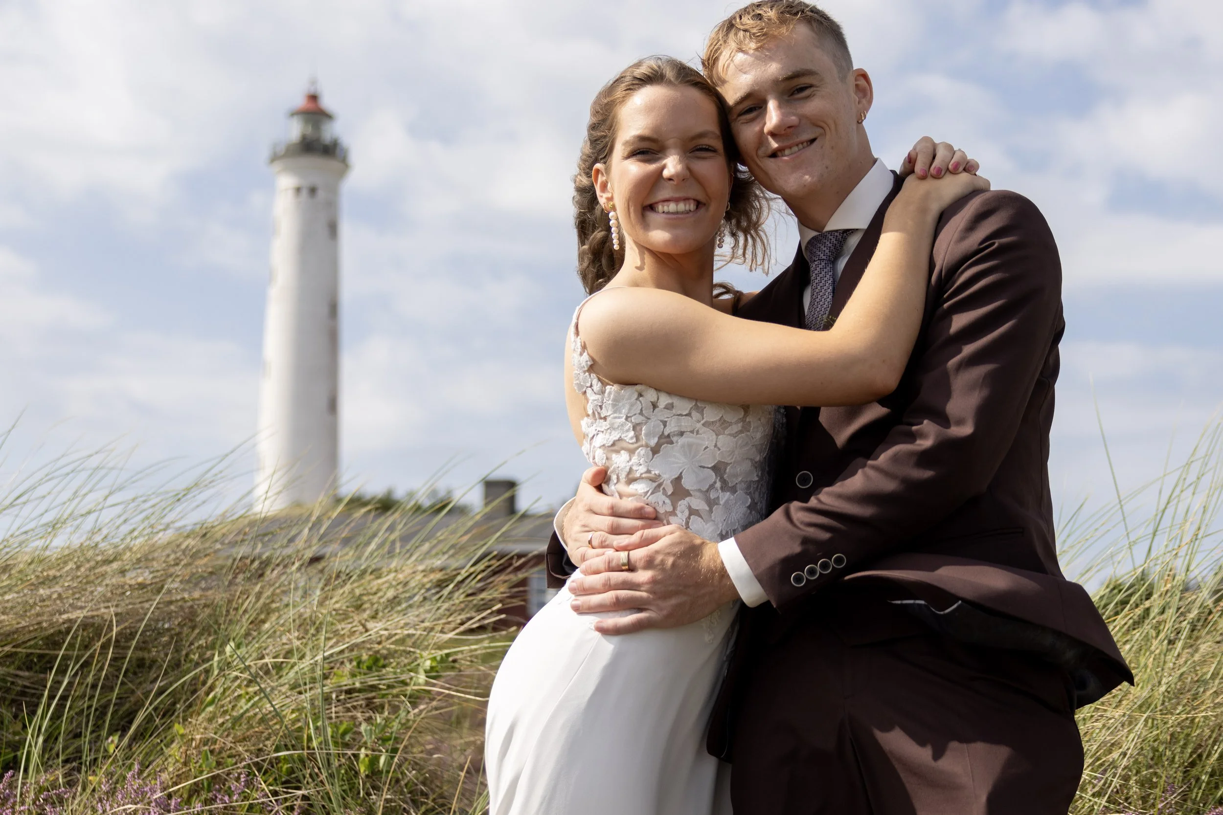 A happy bride and groom embracing outdoors near a lighthouse, smiling at the camera.