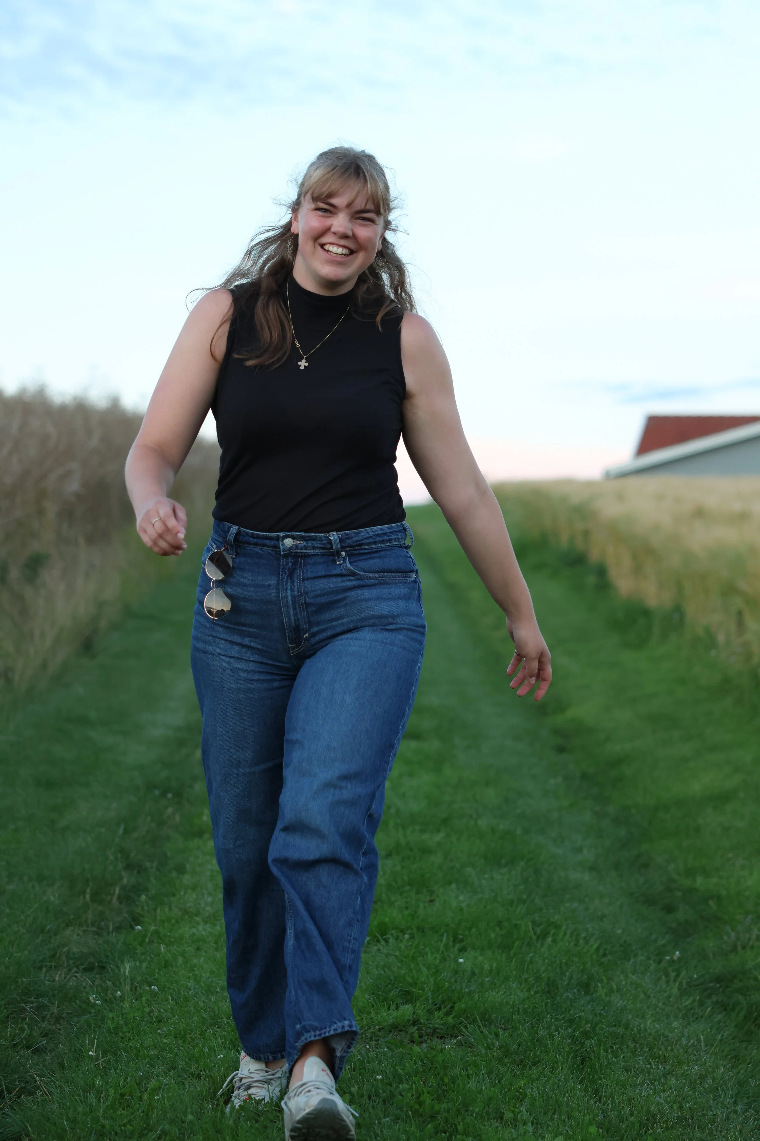 A young woman with long, light brown hair and a wide smile, wearing a sleeveless black top, blue jeans, and white sneakers, walking along a grassy path outdoors with a building and open sky in the background.