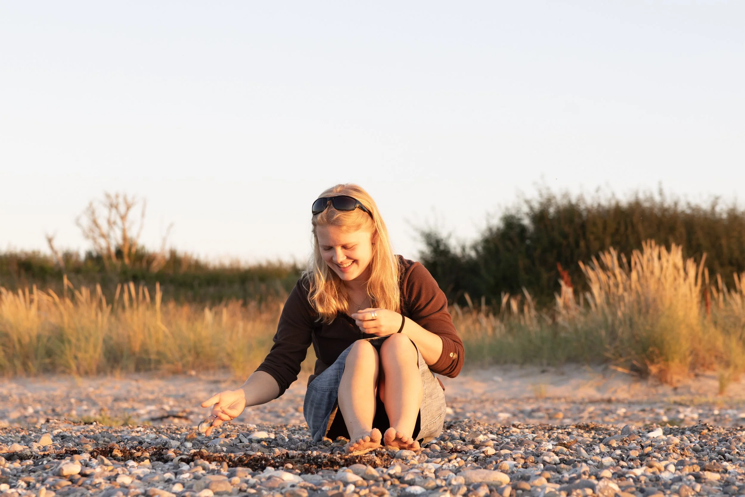 A young woman sitting on a pebbly beach, smiling and reaching out to touch the sand, with grass and bushes in the background during sunset.