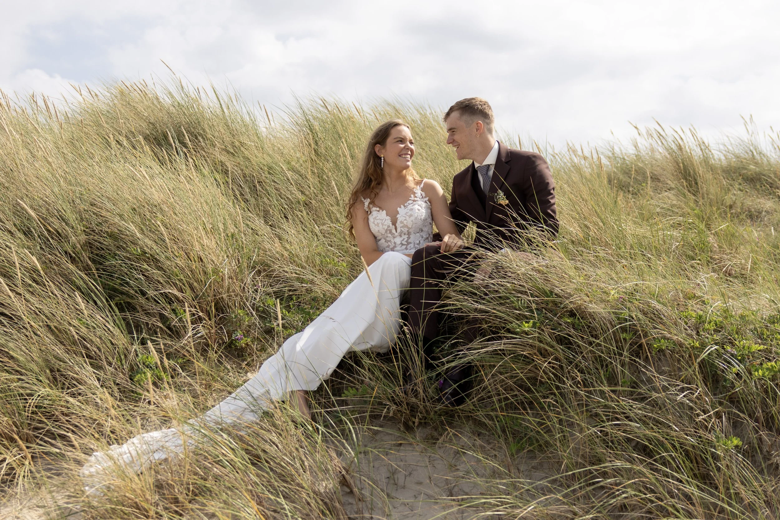 A bride and groom sitting in a grassy dune, smiling and looking at each other on their wedding day.