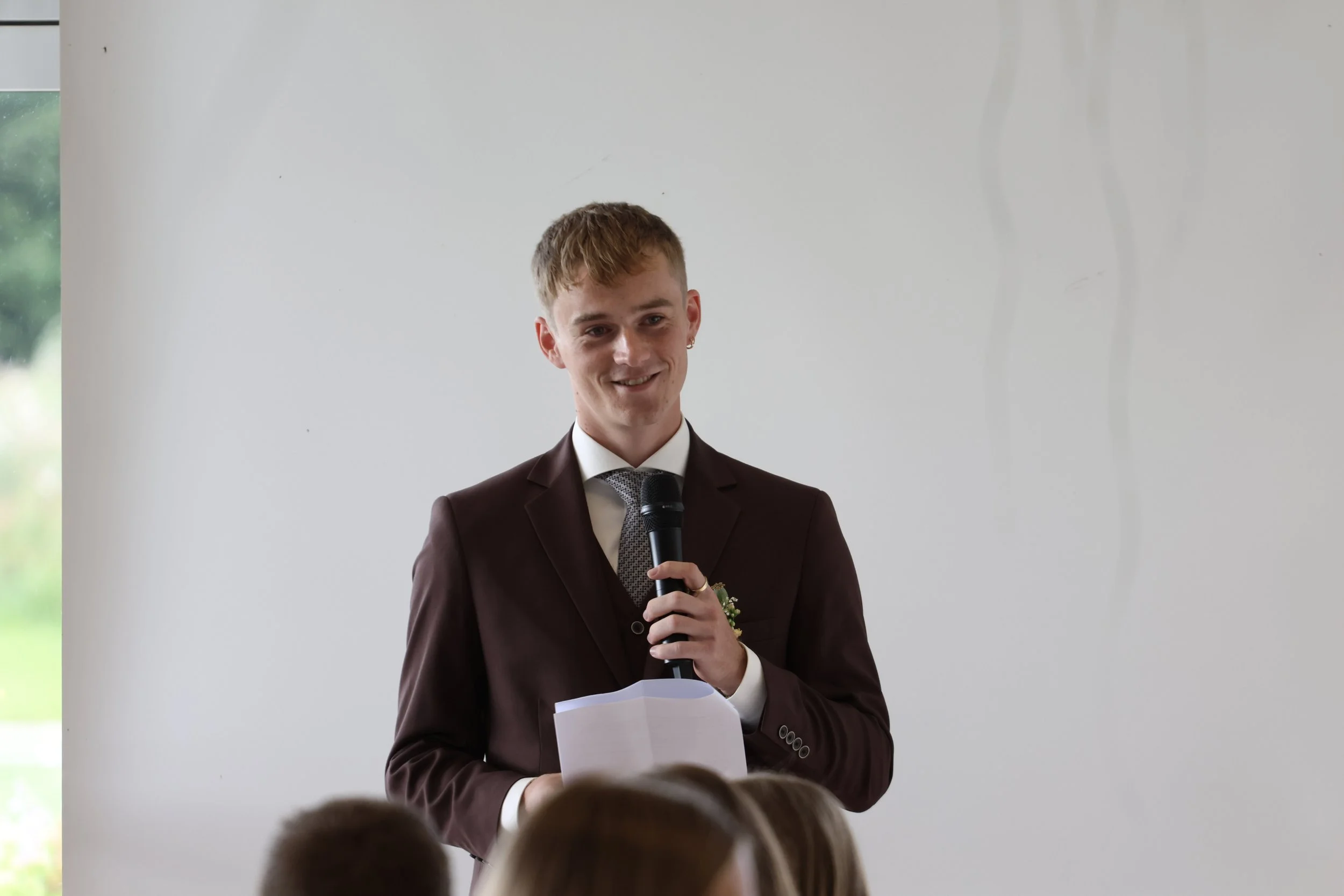 A young man in a brown suit, white shirt, and tie, holding a microphone and a stack of notes, speaking during a formal event, with seated audience members and a white wall in the background.