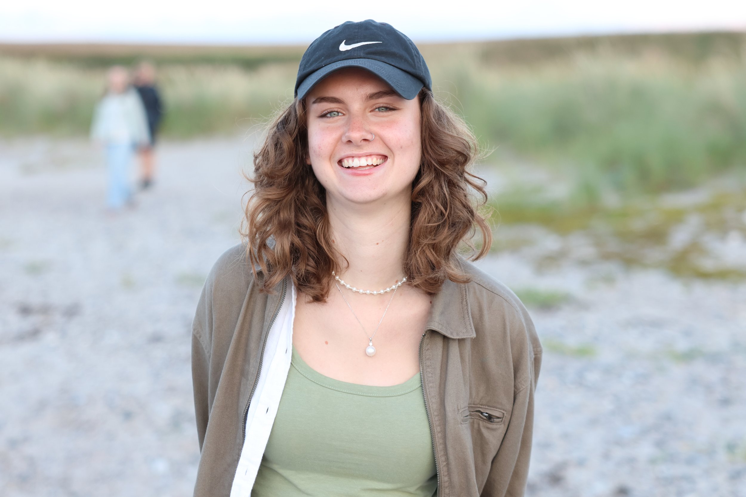 Young woman with curly hair wearing a black Nike cap, a beige jacket, a green top, and pearl jewelry, smiling at the camera outdoors near a beach or riverside.